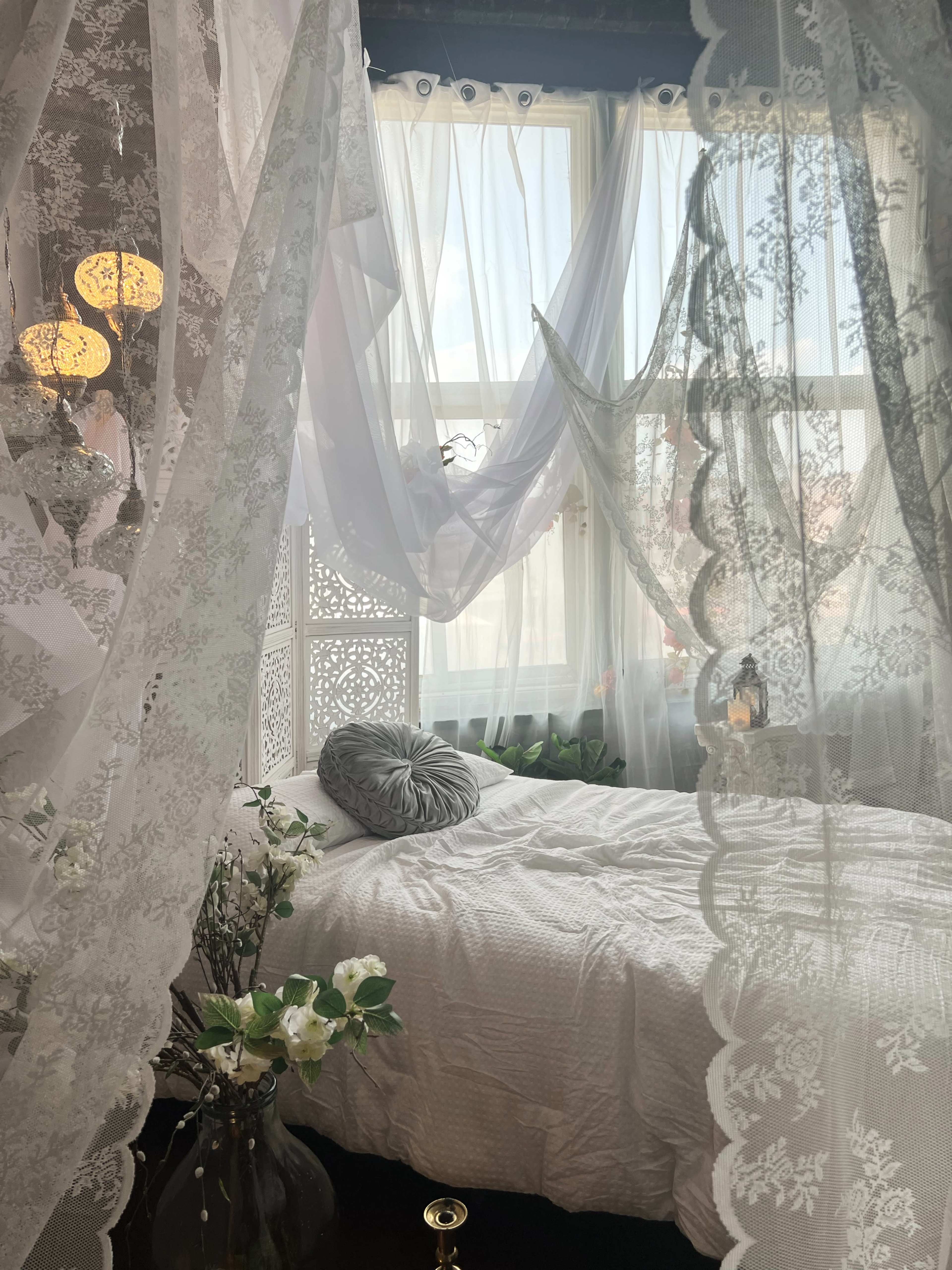 The image shows a cozy bedroom with a white bed draped in sheer lace curtains, surrounded by plants and decorative elements by the window.