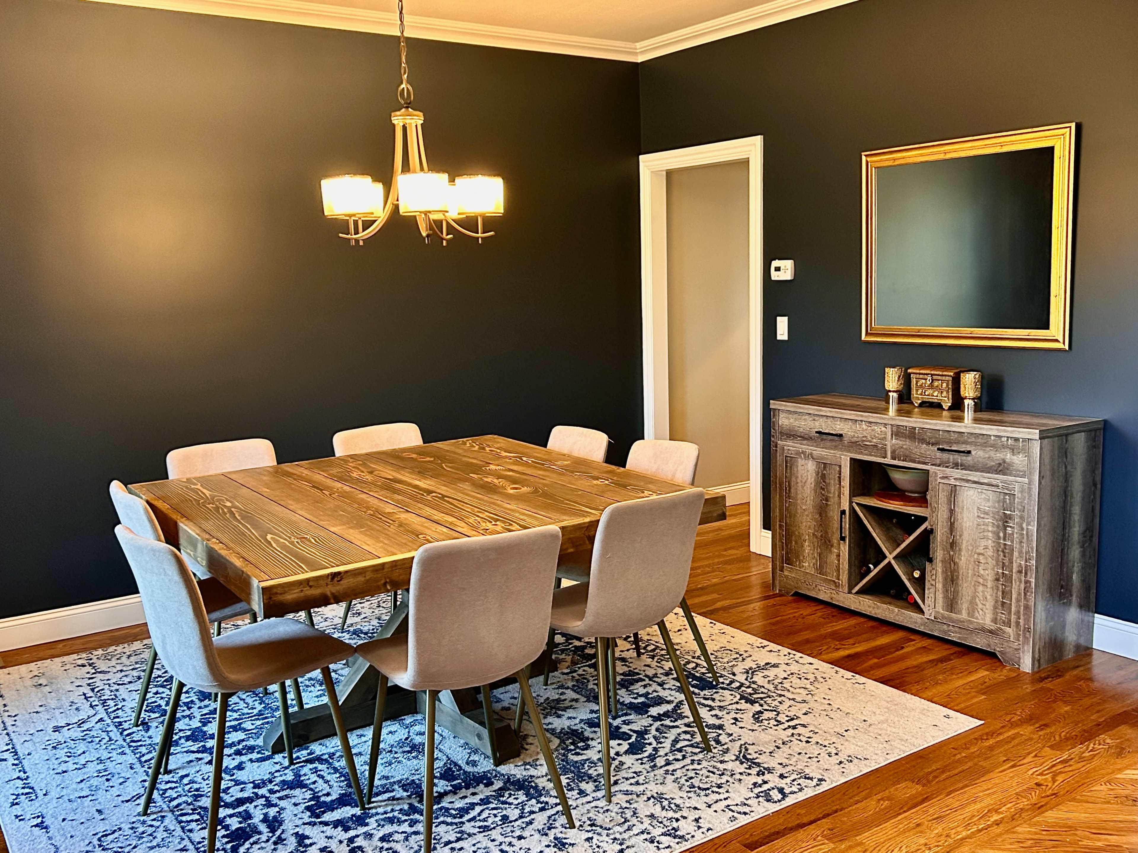 A dining room featuring a wooden table with eight chairs, a sideboard with wine storage, and a decorative mirror on a dark wall.