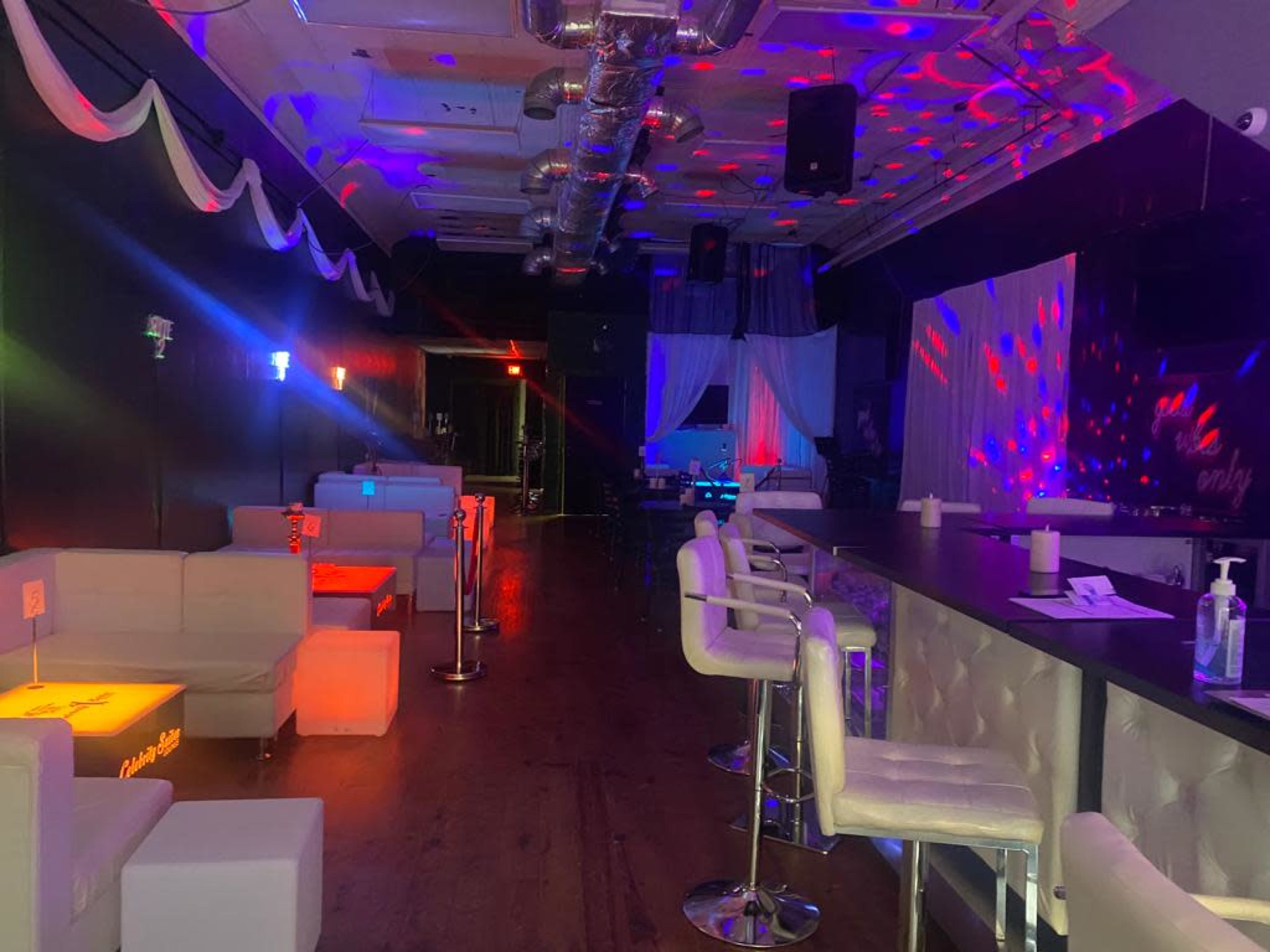 A dimly lit bar area with white lounge seating, illuminated by colorful stage lights and decorations.