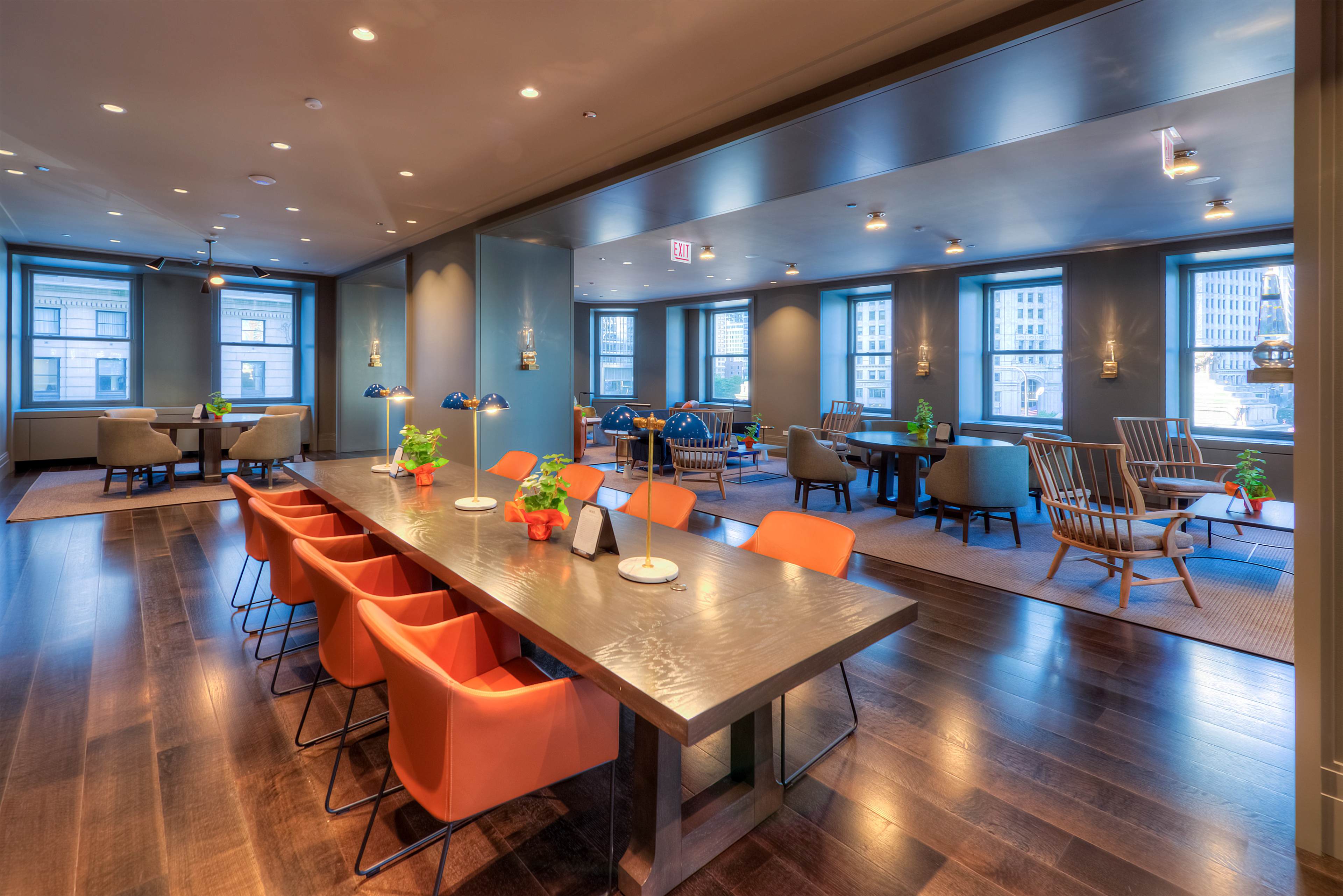A modern room with a long dining table surrounded by orange chairs, alongside seating areas with plants and large windows letting in natural light.