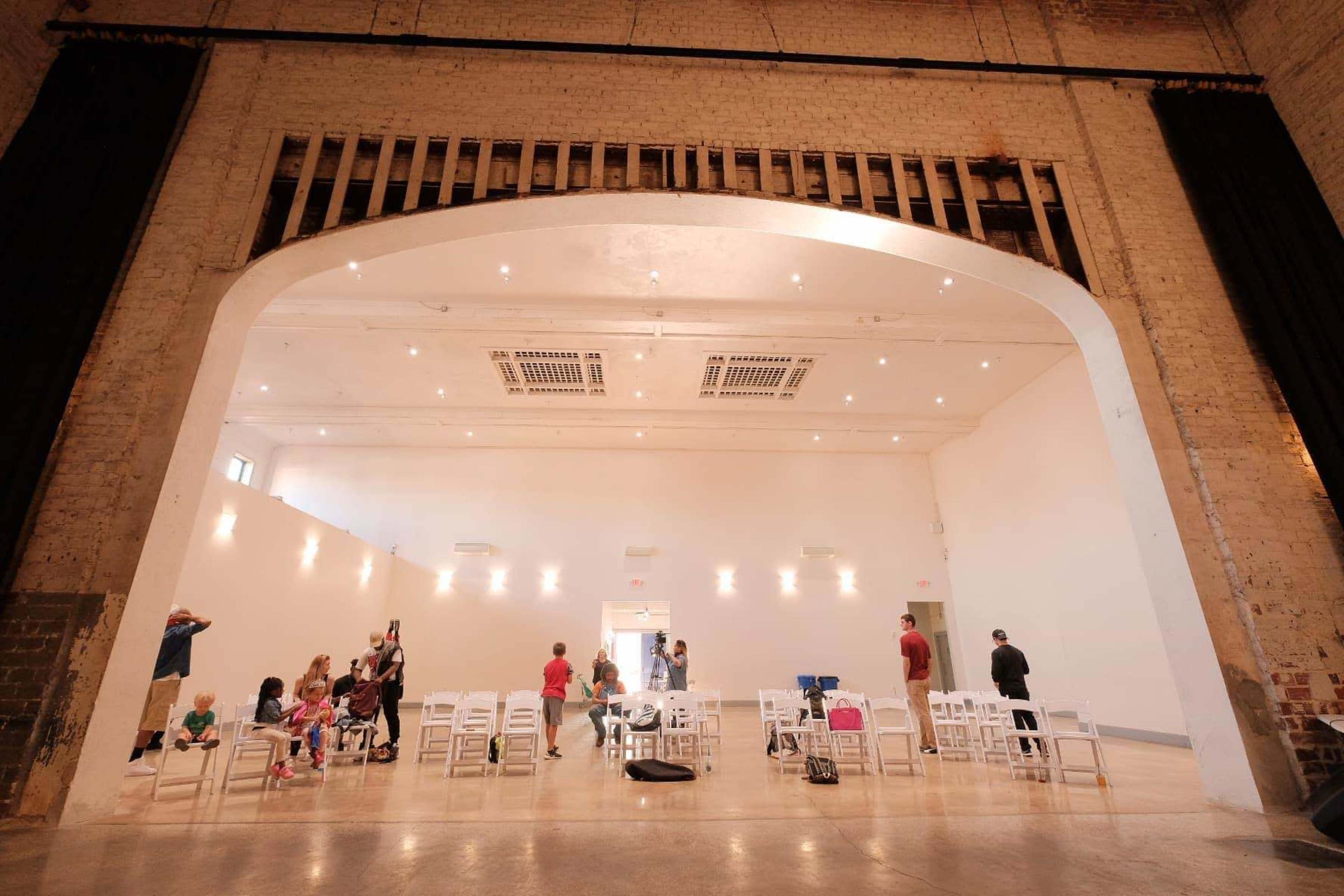 A spacious, brightly lit interior of a hall with several white chairs arranged for an event, while people are moving around and interacting.
