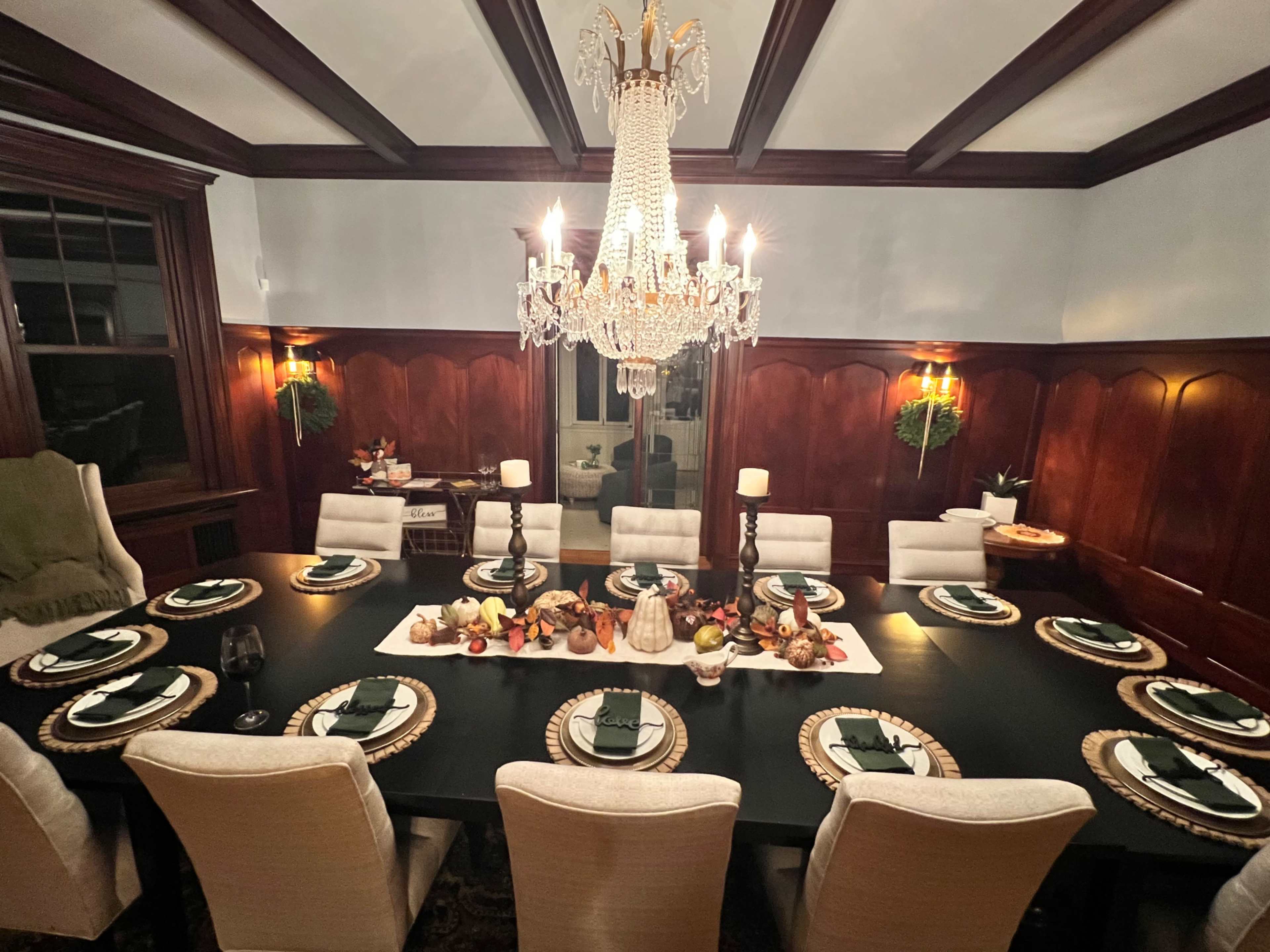 A formal dining room is set with a long table adorned with seasonal decorations, plates, and candles, under a sparkling chandelier.