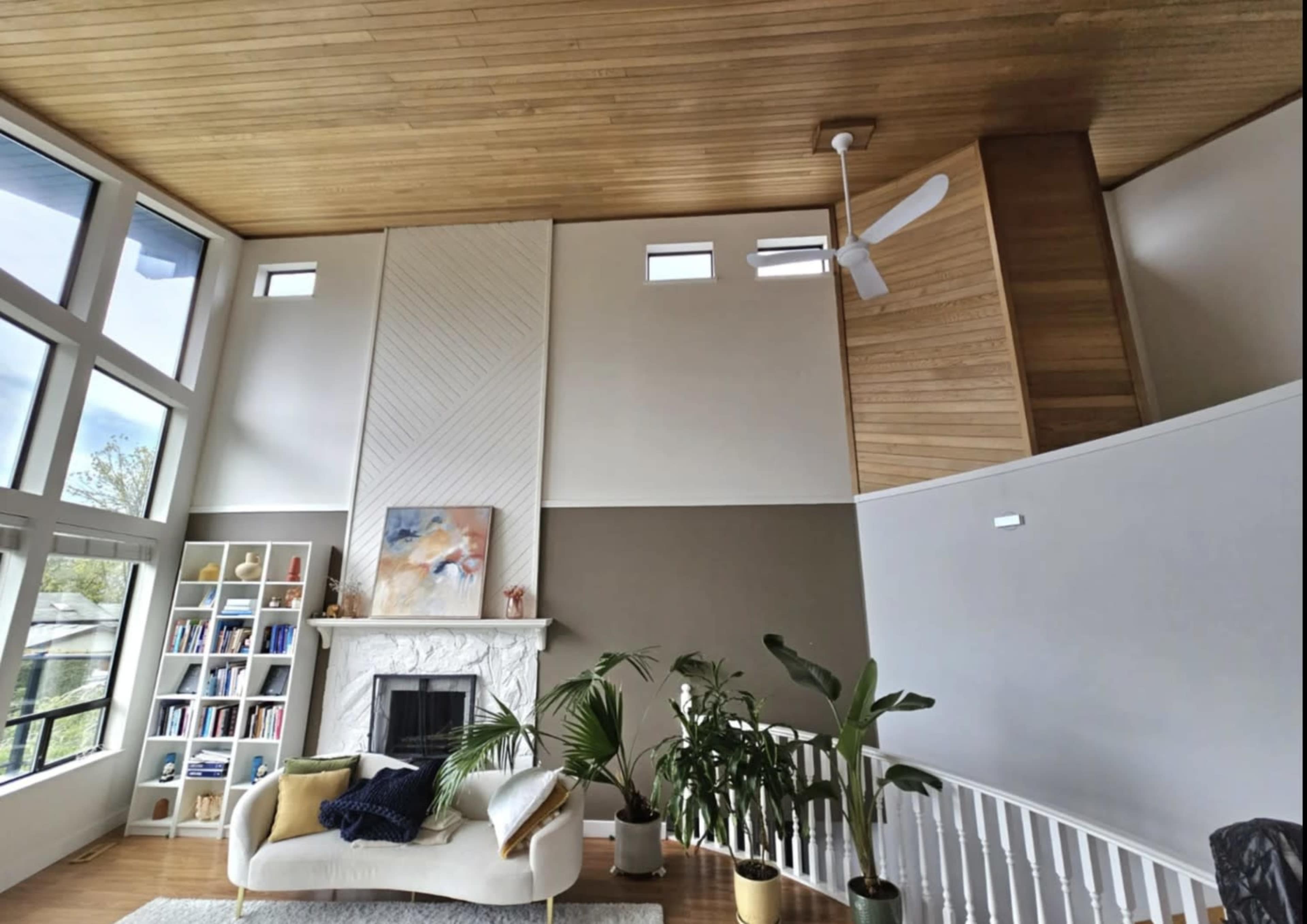 The image shows a spacious living room with large windows, a white stone fireplace, a light-colored sofa, and several indoor plants.