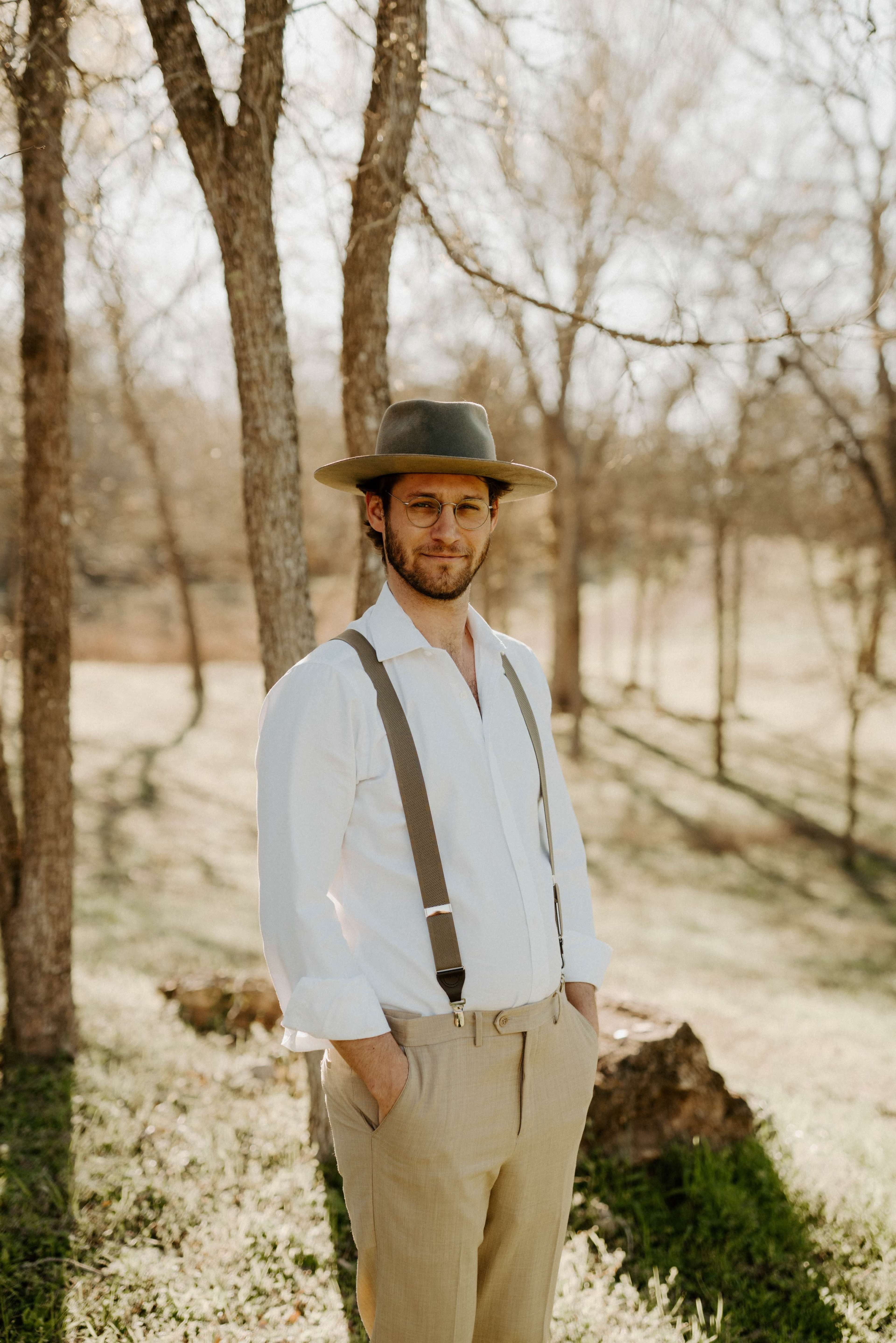 A man in a white shirt and suspenders stands outdoors in a wooded area with trees in the background.