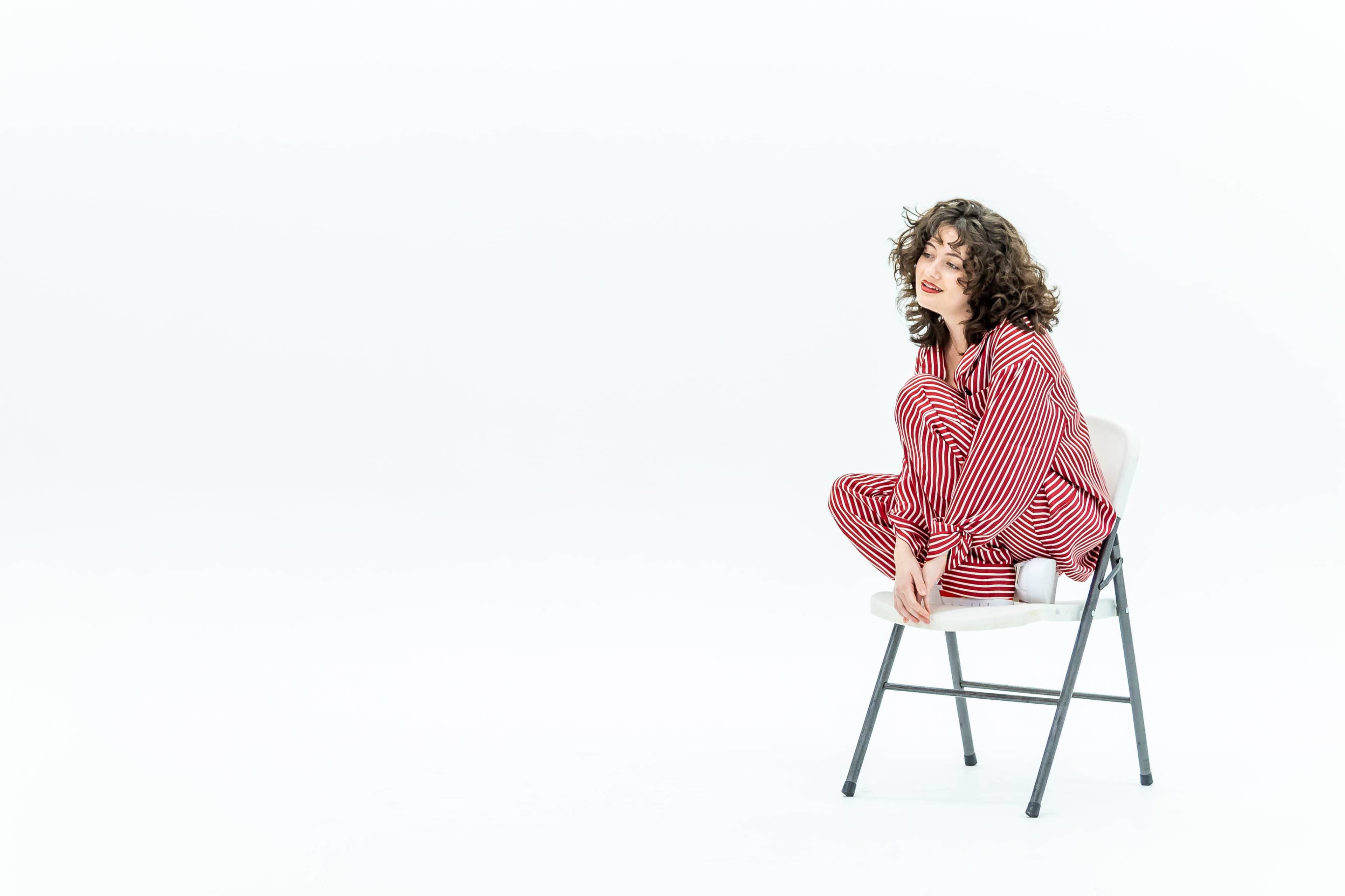 A person with curly hair sits on a chair, wearing red and white striped pajamas in a minimalist, white background setting.