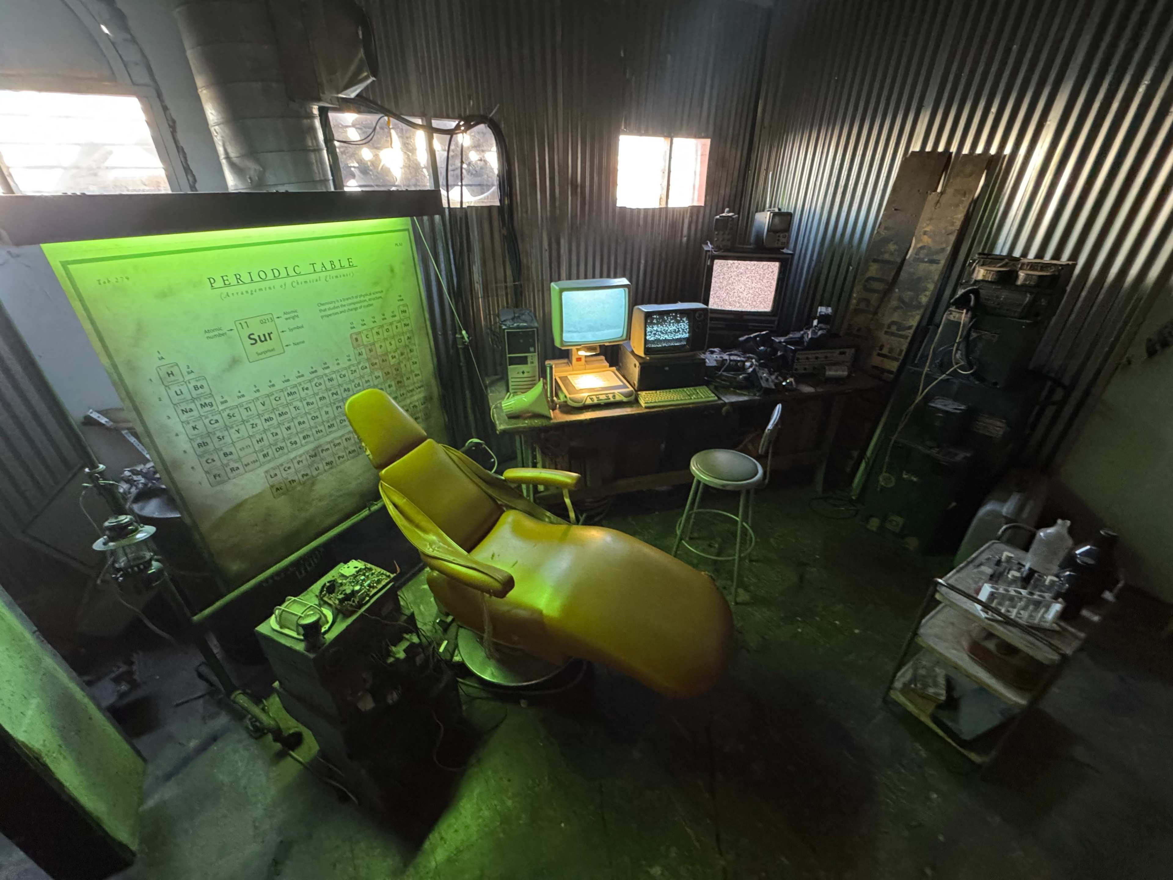 The image shows a dimly lit room with a yellow dental chair, a vintage computer setup, and various equipment against a corrugated metal wall.