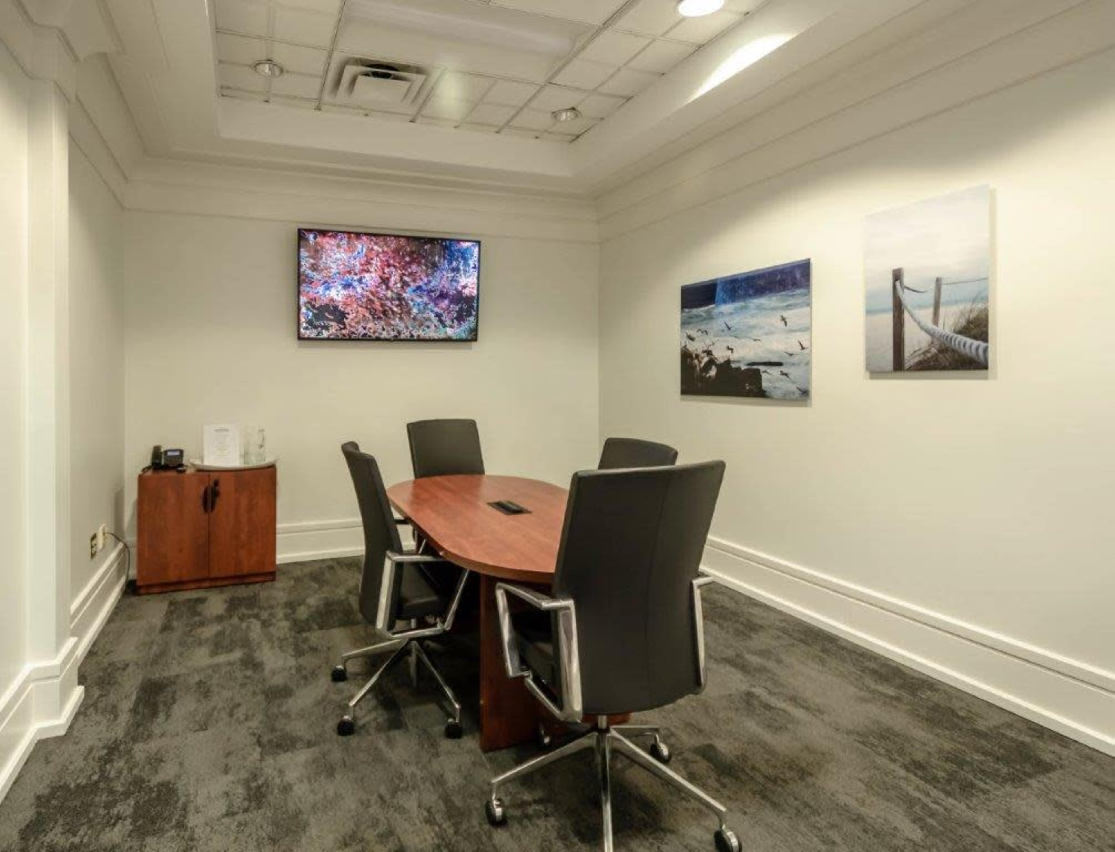 A small conference room with a wooden table and four black chairs, featuring a TV mounted on the wall and two framed pictures.