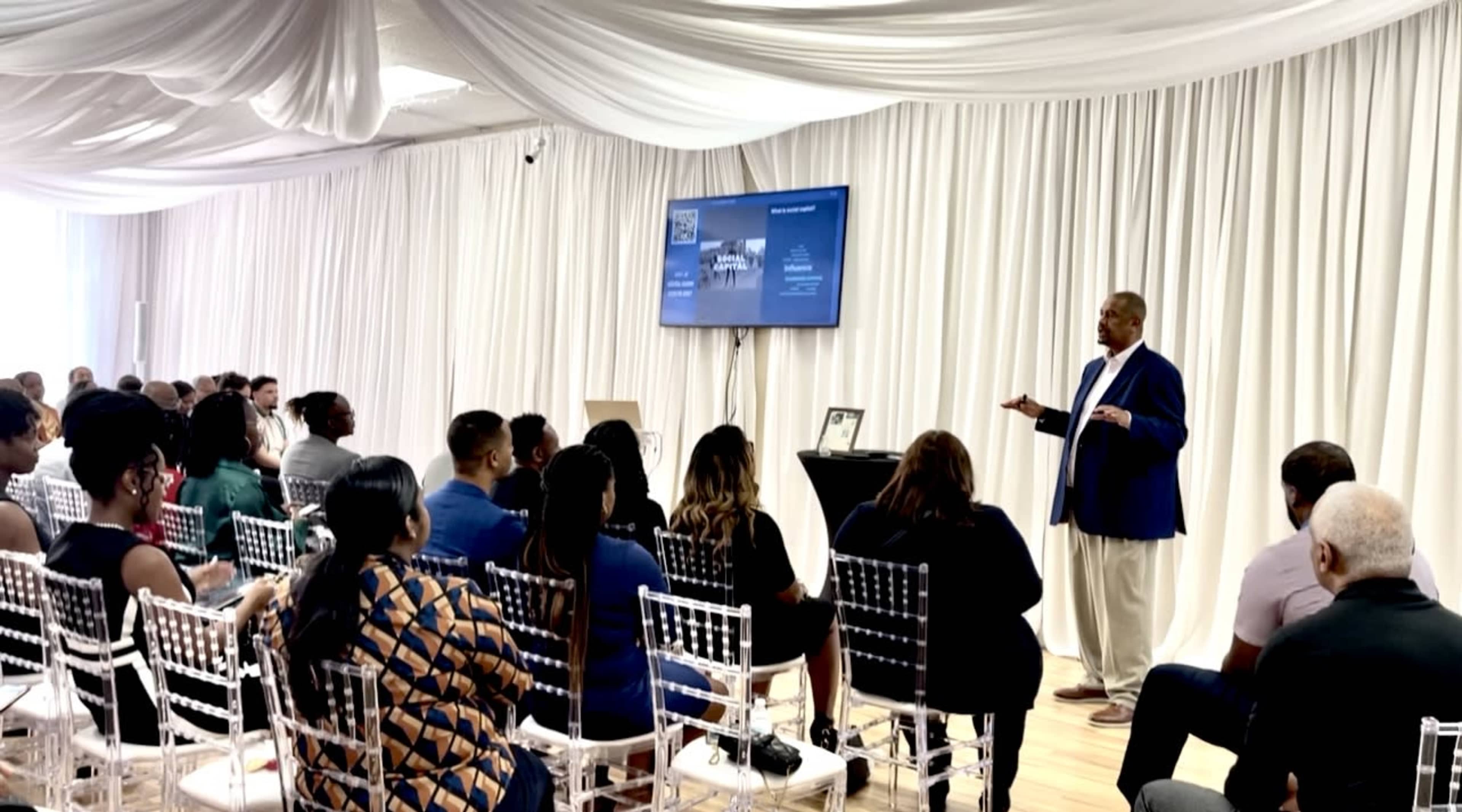 A speaker presents to an audience seated in a well-lit room with draped curtains and a screen displaying information.