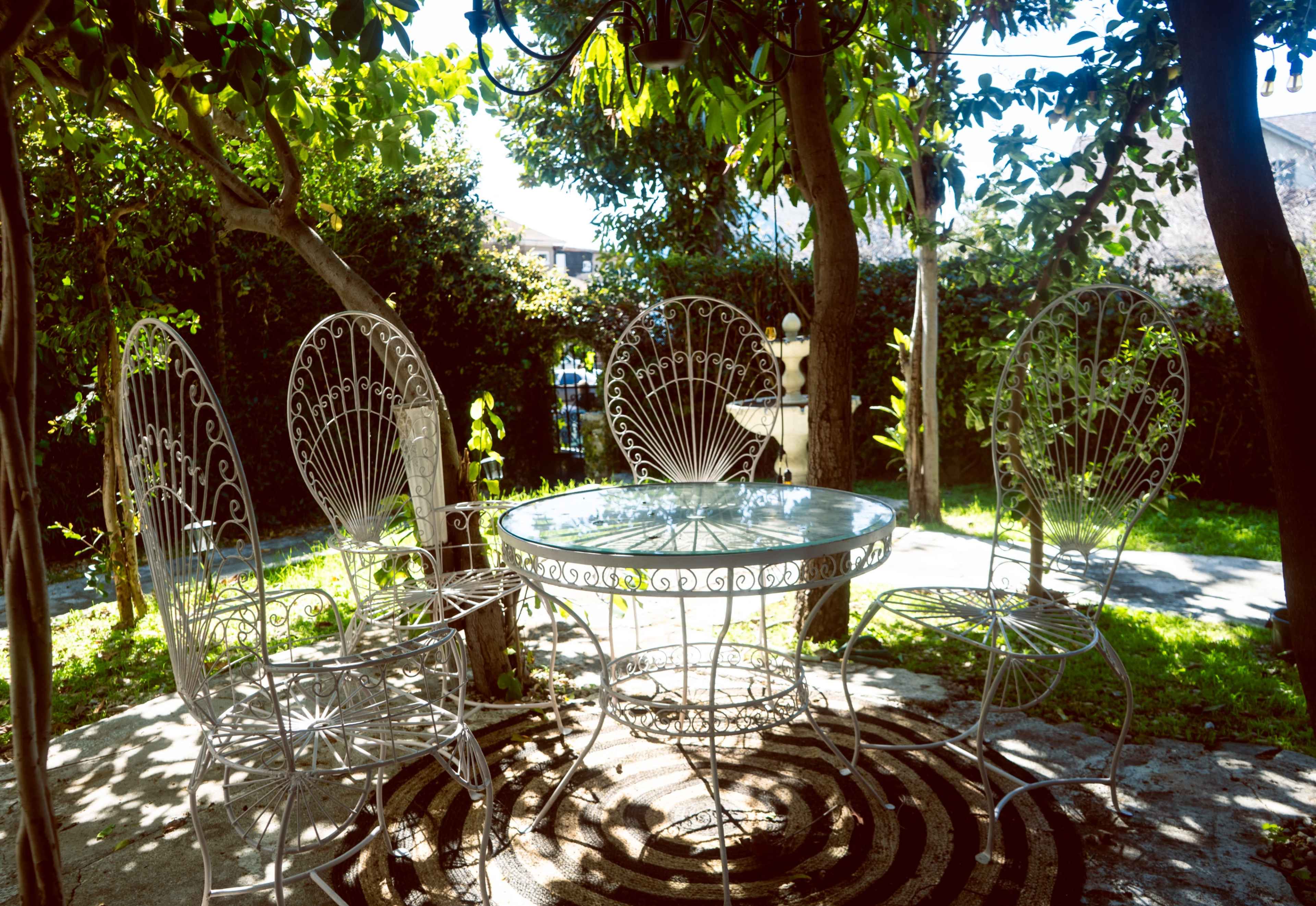 A round glass table surrounded by four ornate metal chairs is situated under leafy trees in a garden space.