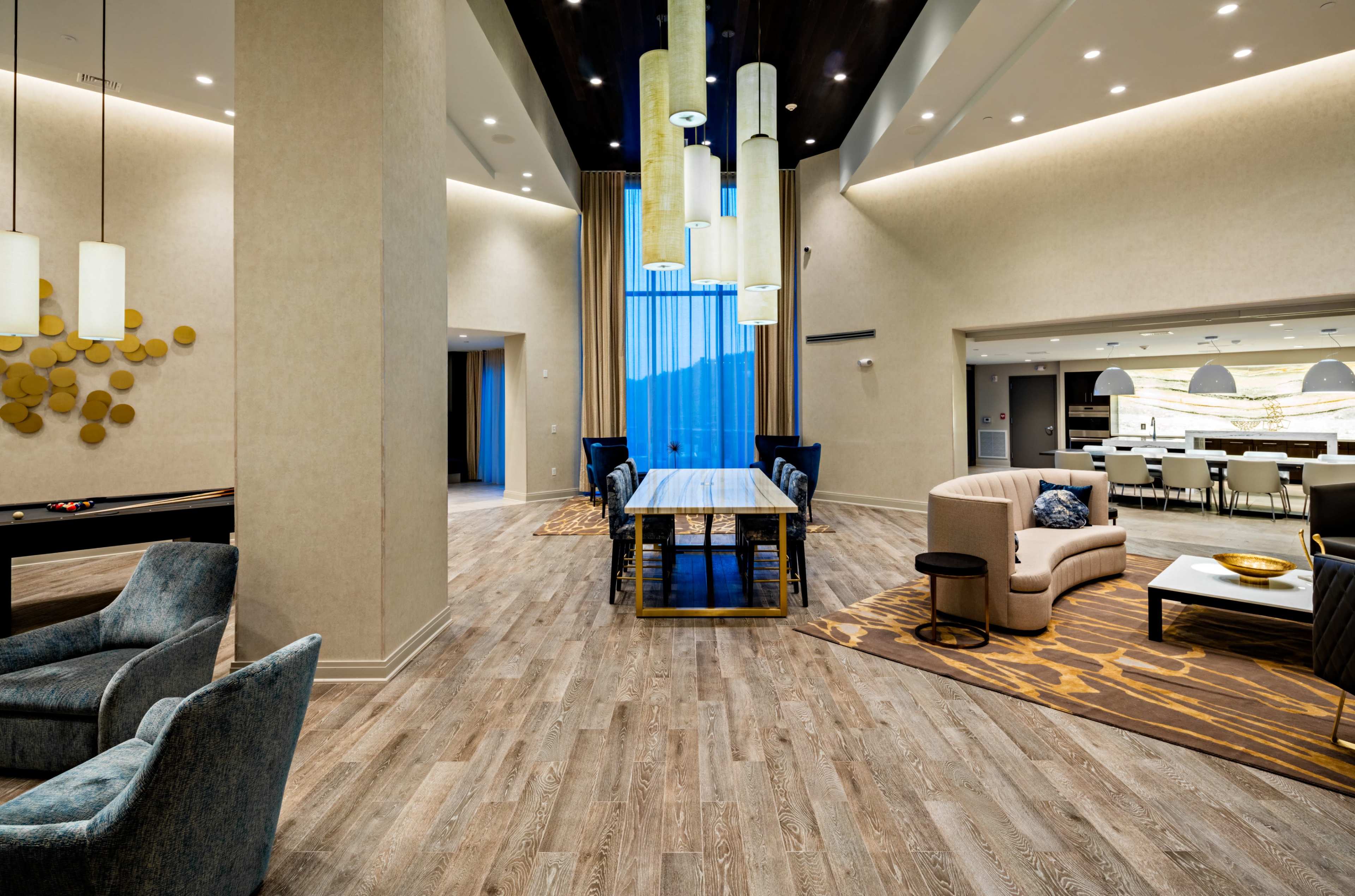 The image shows a modern interior space with a large wooden table surrounded by chairs, illuminated by pendant lights, and featuring a mix of seating areas and large windows.