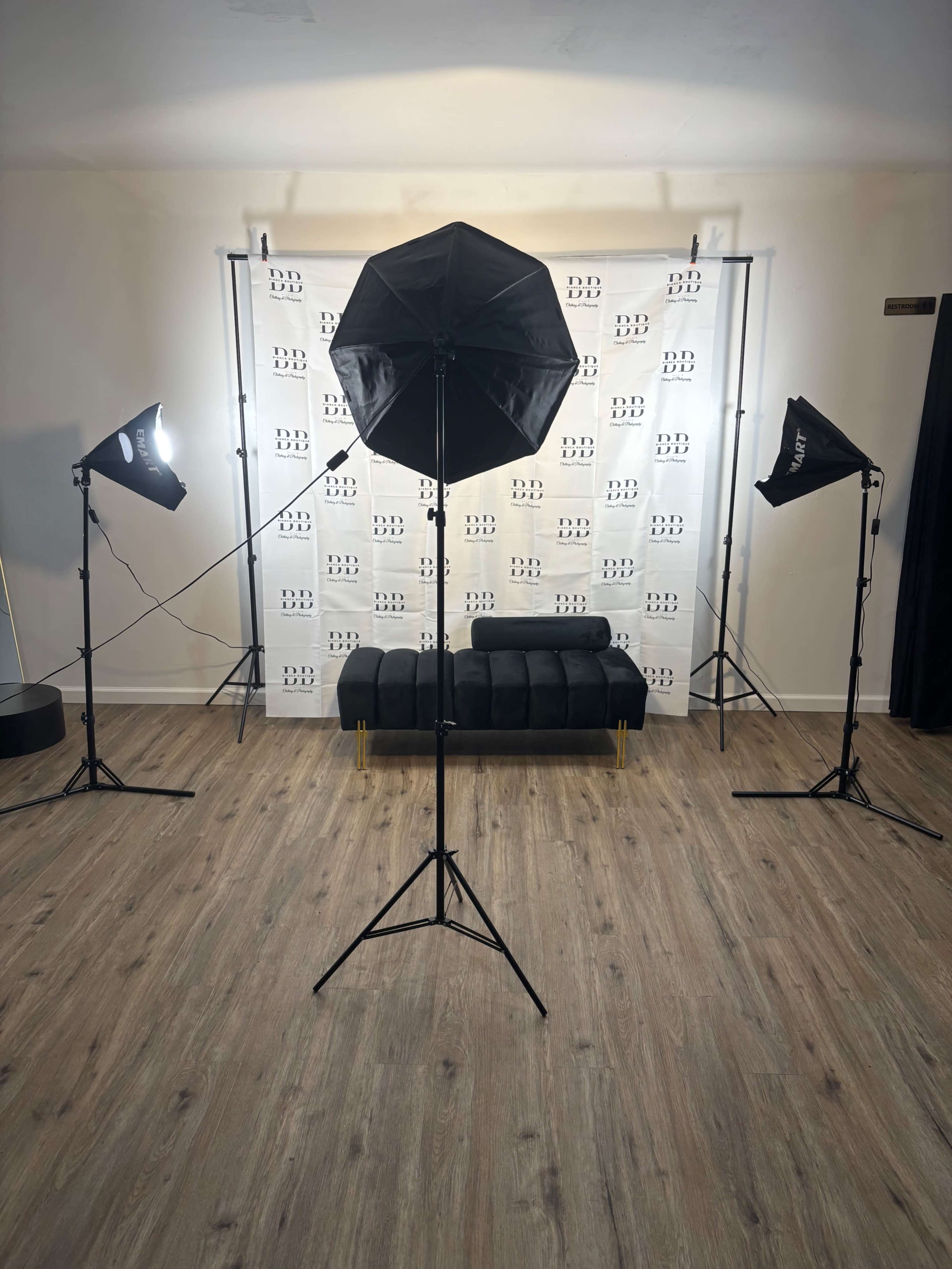 A photography studio setup features a black couch in front of a branded backdrop, illuminated by multiple light sources on tripods.
