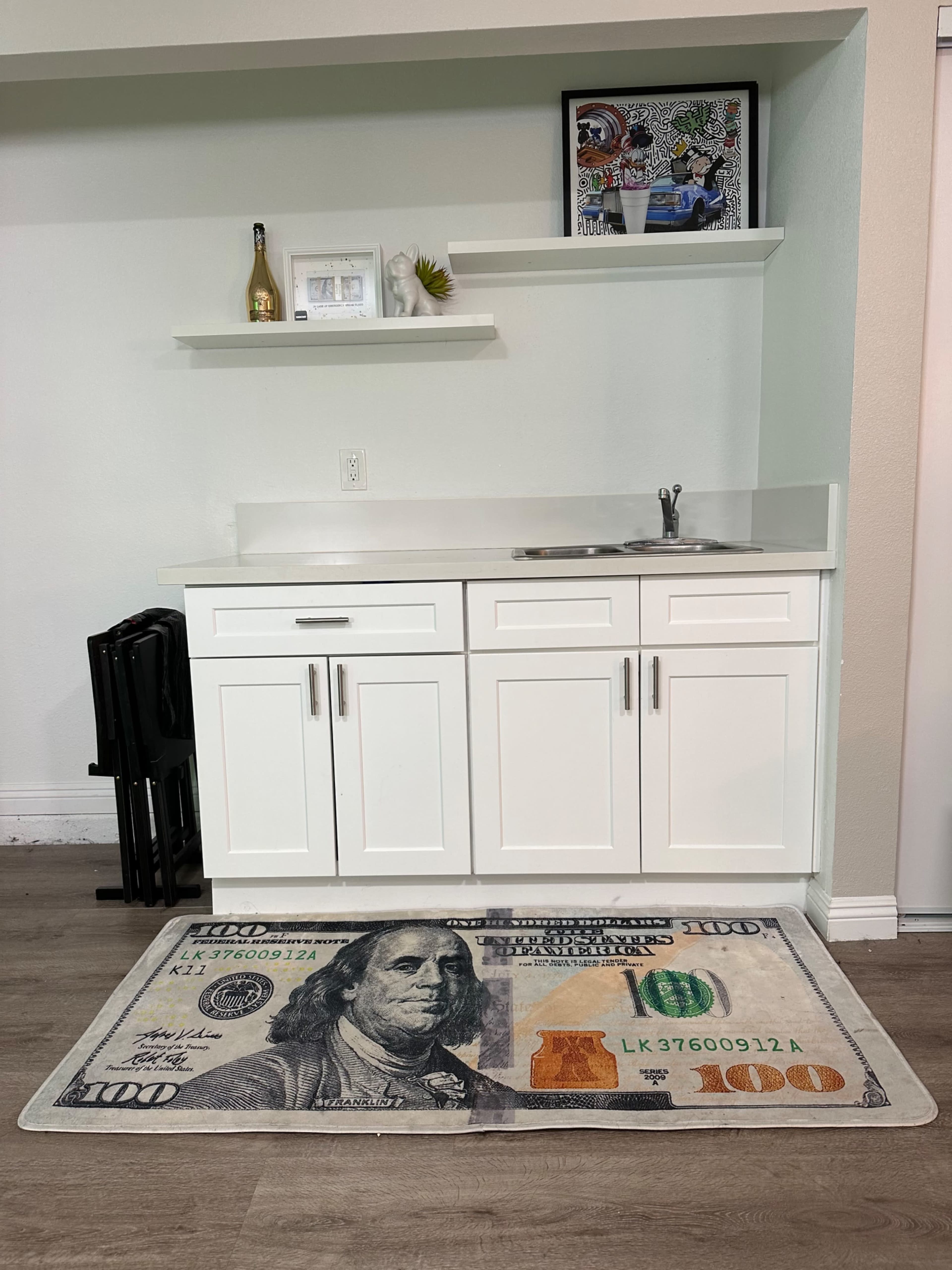 A kitchen area with white cabinets, a small sink, and a large rug designed to resemble a one hundred dollar bill.