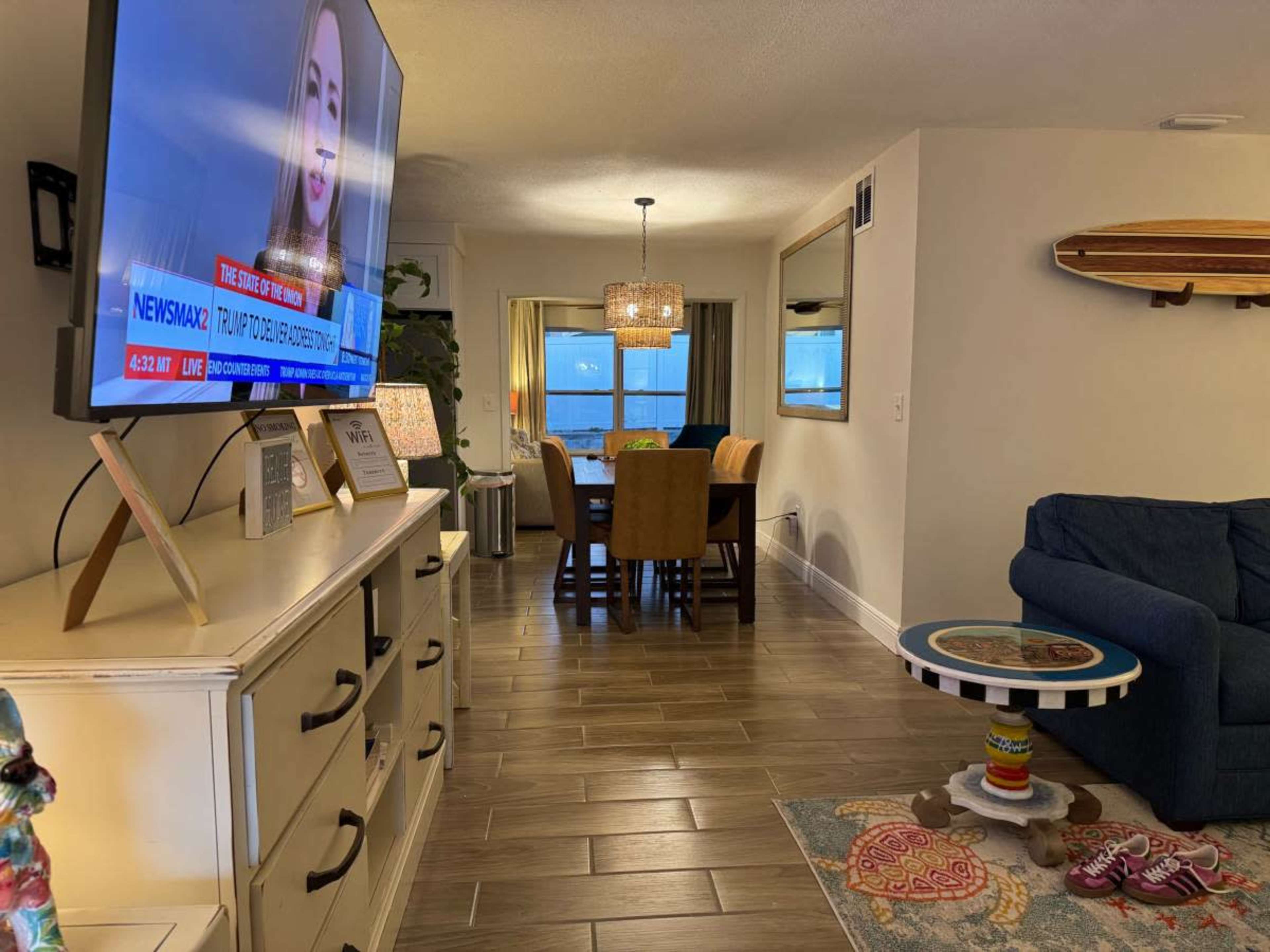 The image shows a living room with a blue couch, a television displaying a news broadcast, a dining area in the background, and decorative items like a colorful rug and a coffee table.