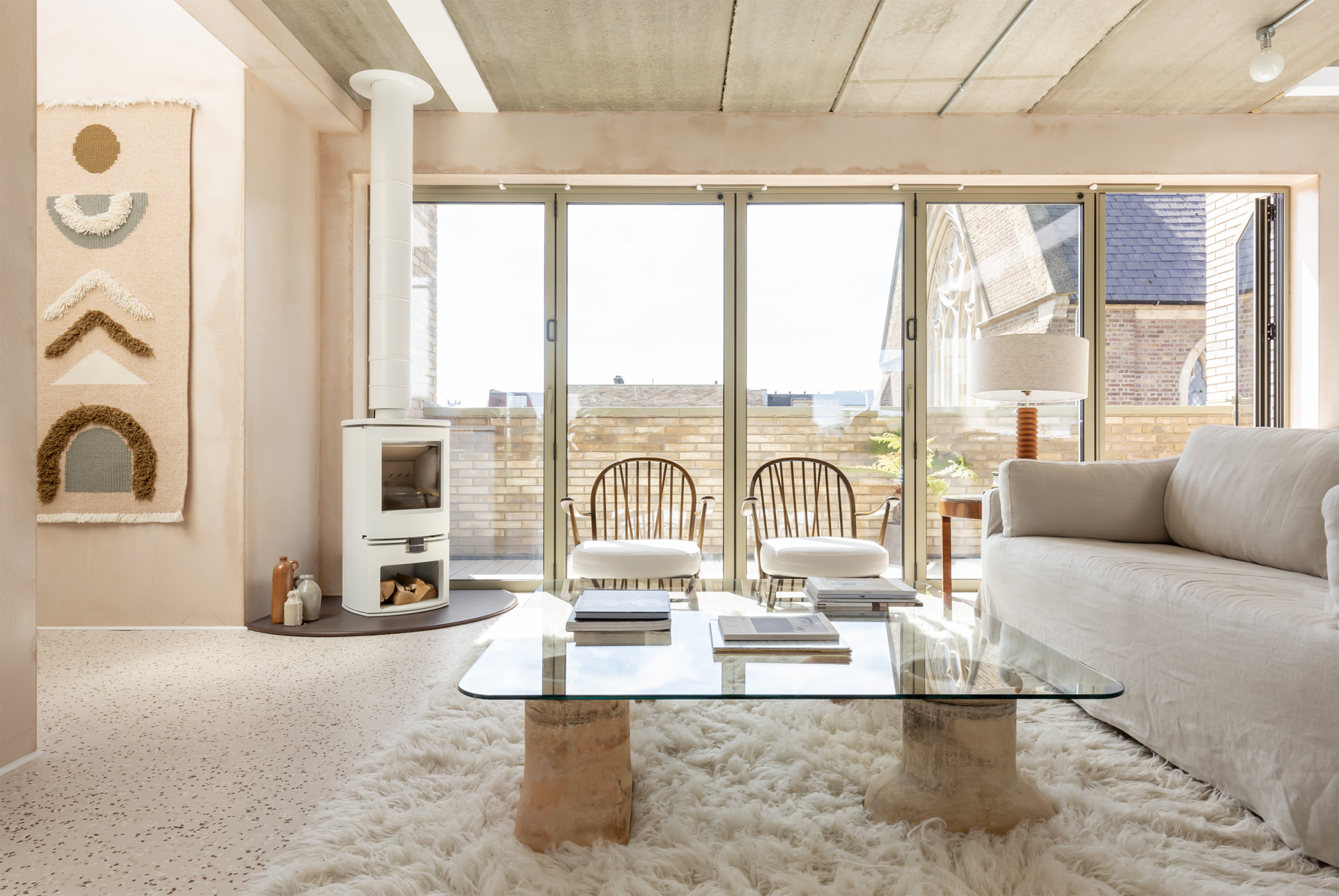 A modern living room features a glass coffee table, two rattan chairs, a white sofa, and a cozy interior with large windows letting in natural light.