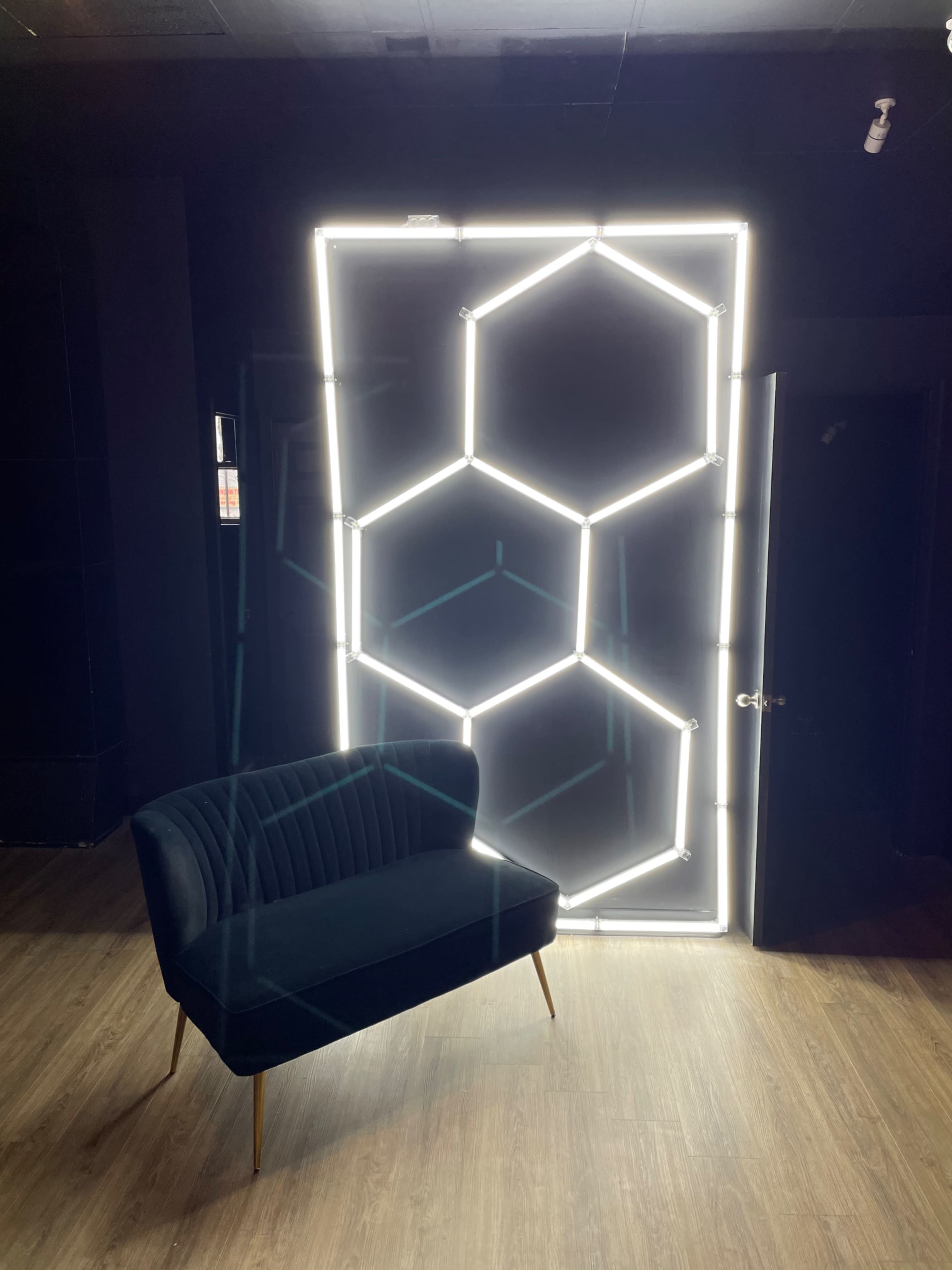 A modern black velvet sofa is positioned in front of a glowing hexagon-patterned light installation against a dark wall.