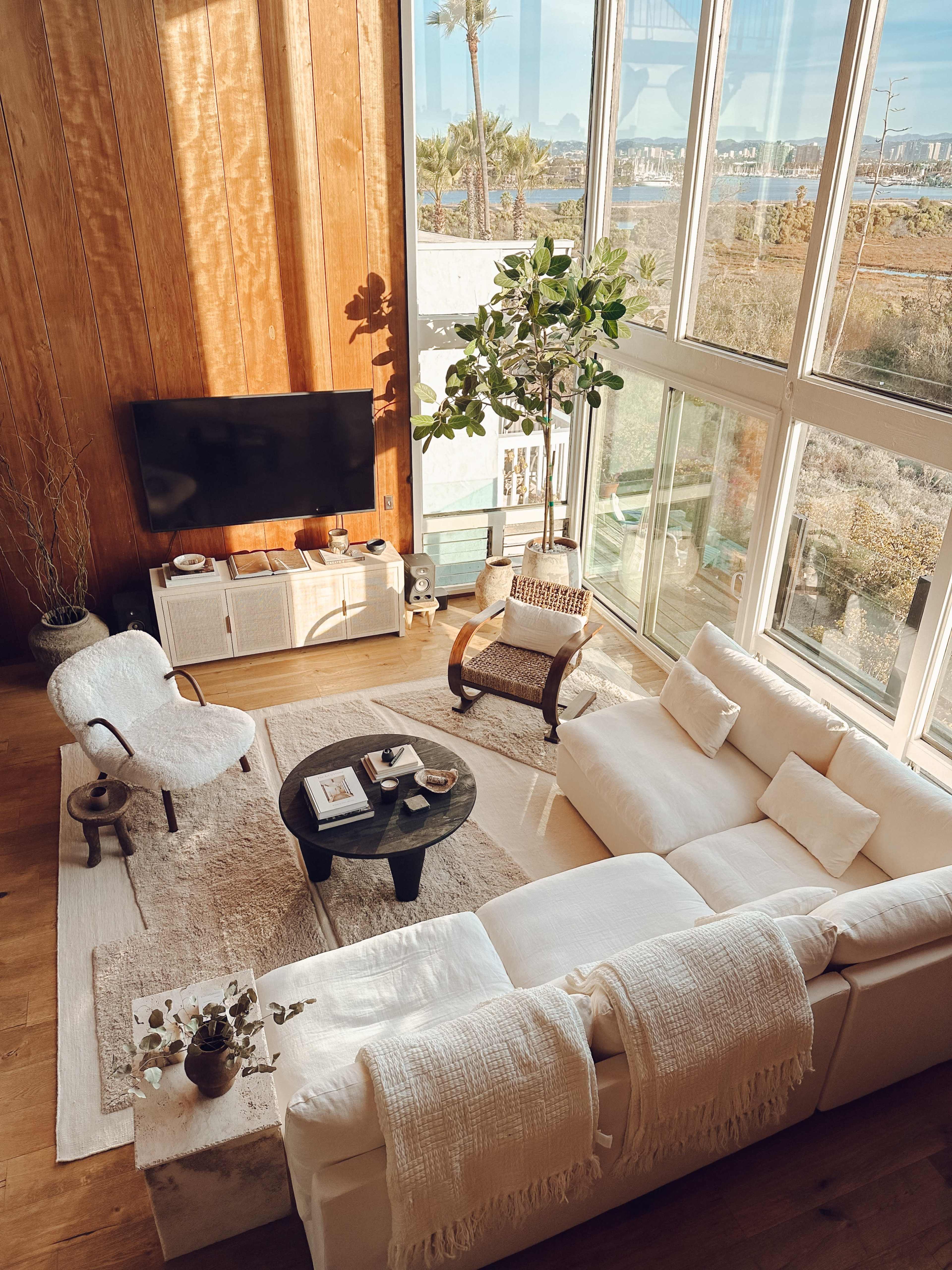 A spacious, sunlit living room with large windows, a white sectional sofa, a round black coffee table, and a natural wood wall.