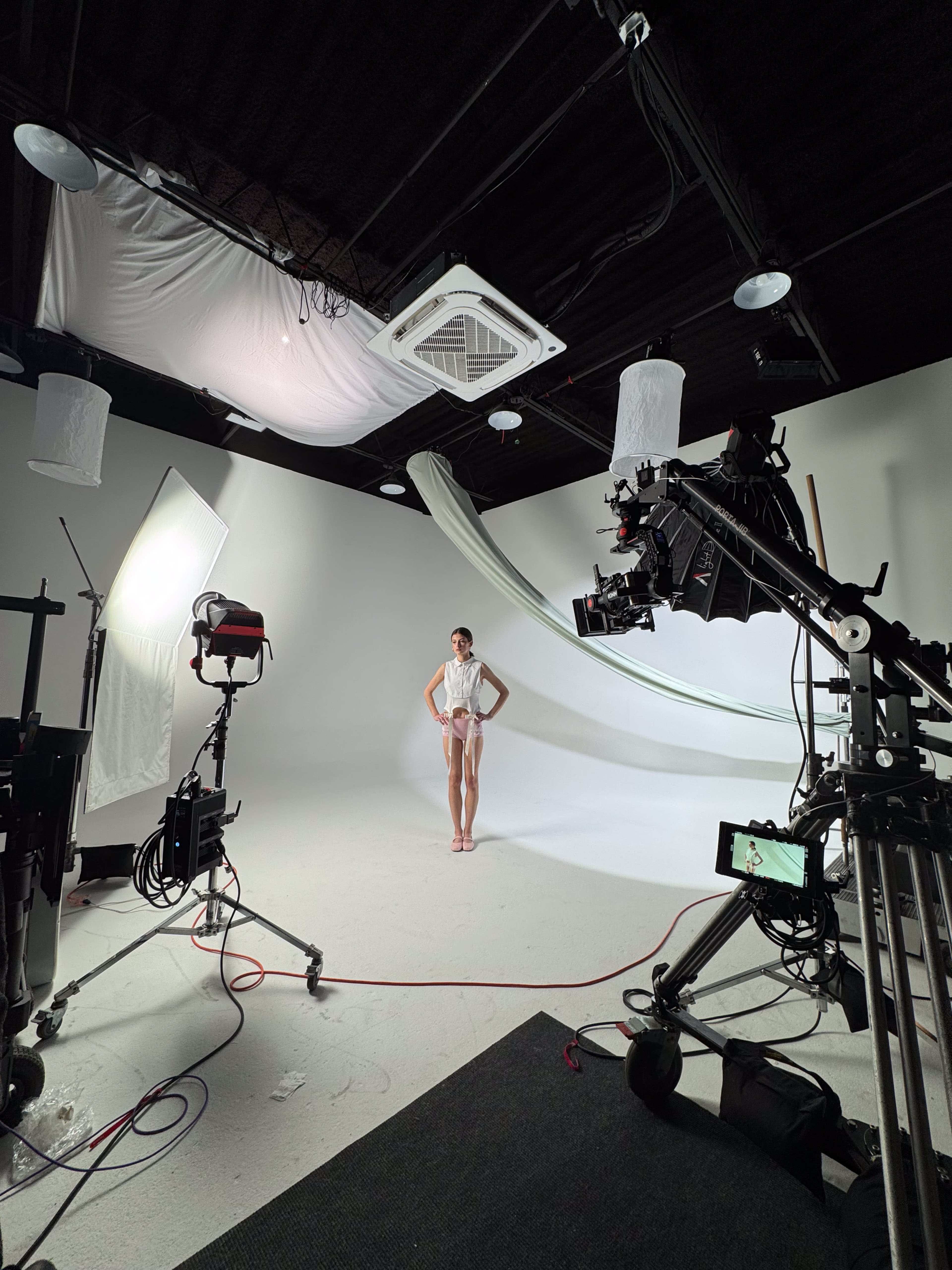 A model poses in a studio with a white backdrop surrounded by lighting equipment and camera setups.