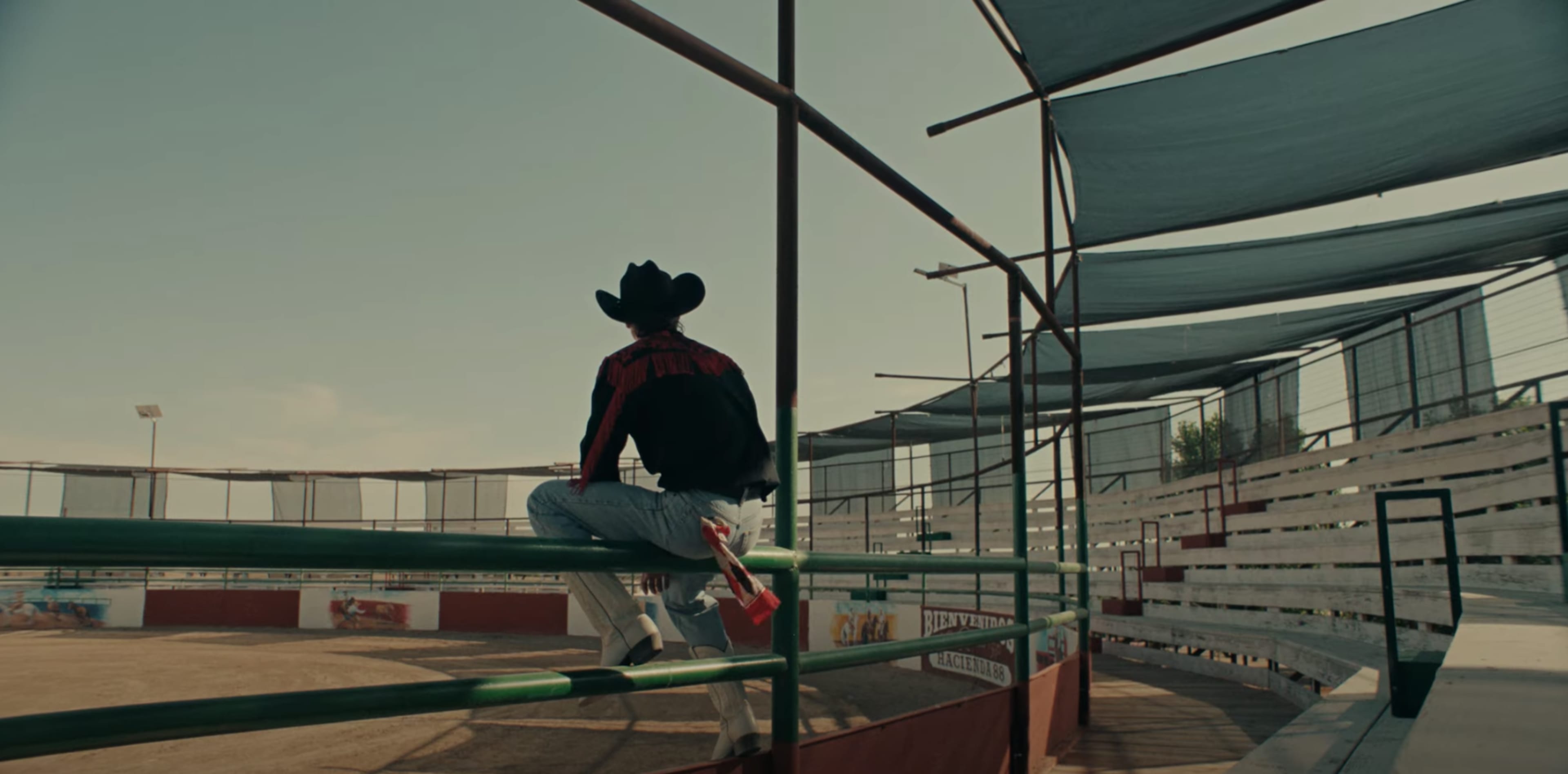 A man in a cowboy hat sits on a railing overlooking an empty arena.
