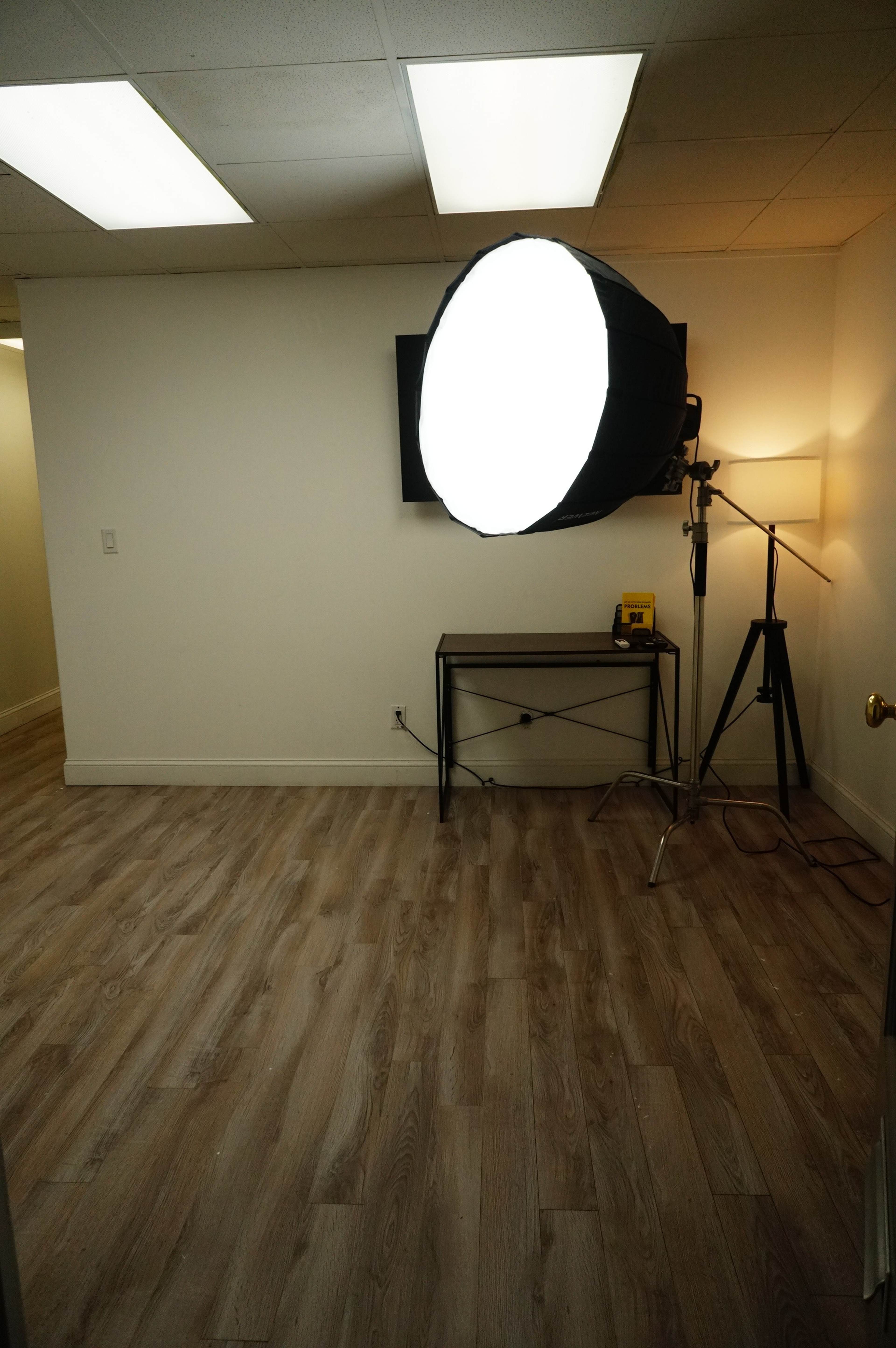 A well-lit room with a large softbox light set up next to a lamp, a television on the wall, and a simple metal table.