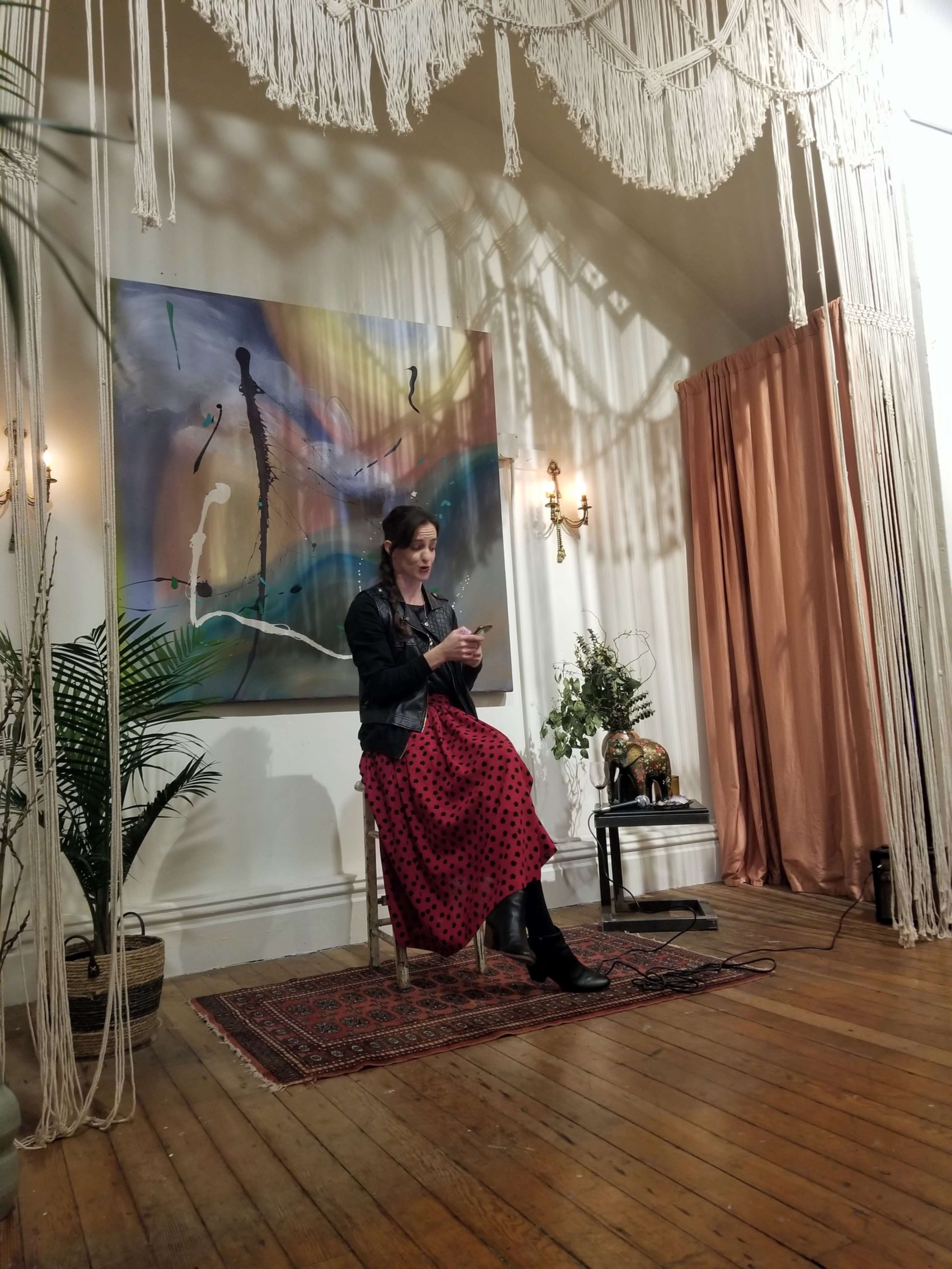 A woman in a black jacket and polka dot skirt sits on a wooden chair, looking at her phone against a backdrop of draped fabric and a large colorful painting.
