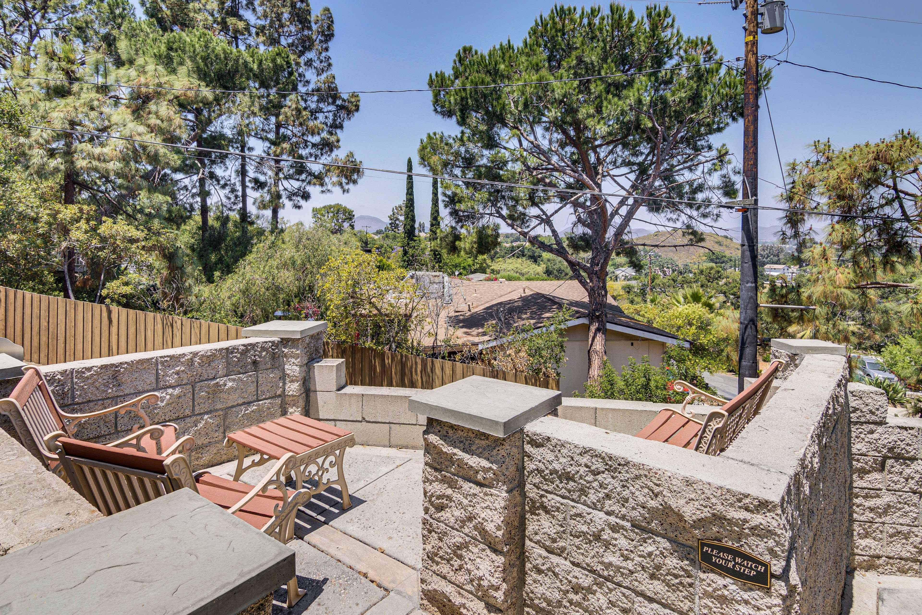 The image shows a stone patio area with metal outdoor furniture, surrounded by trees and overlooking a neighborhood with houses in the distance.