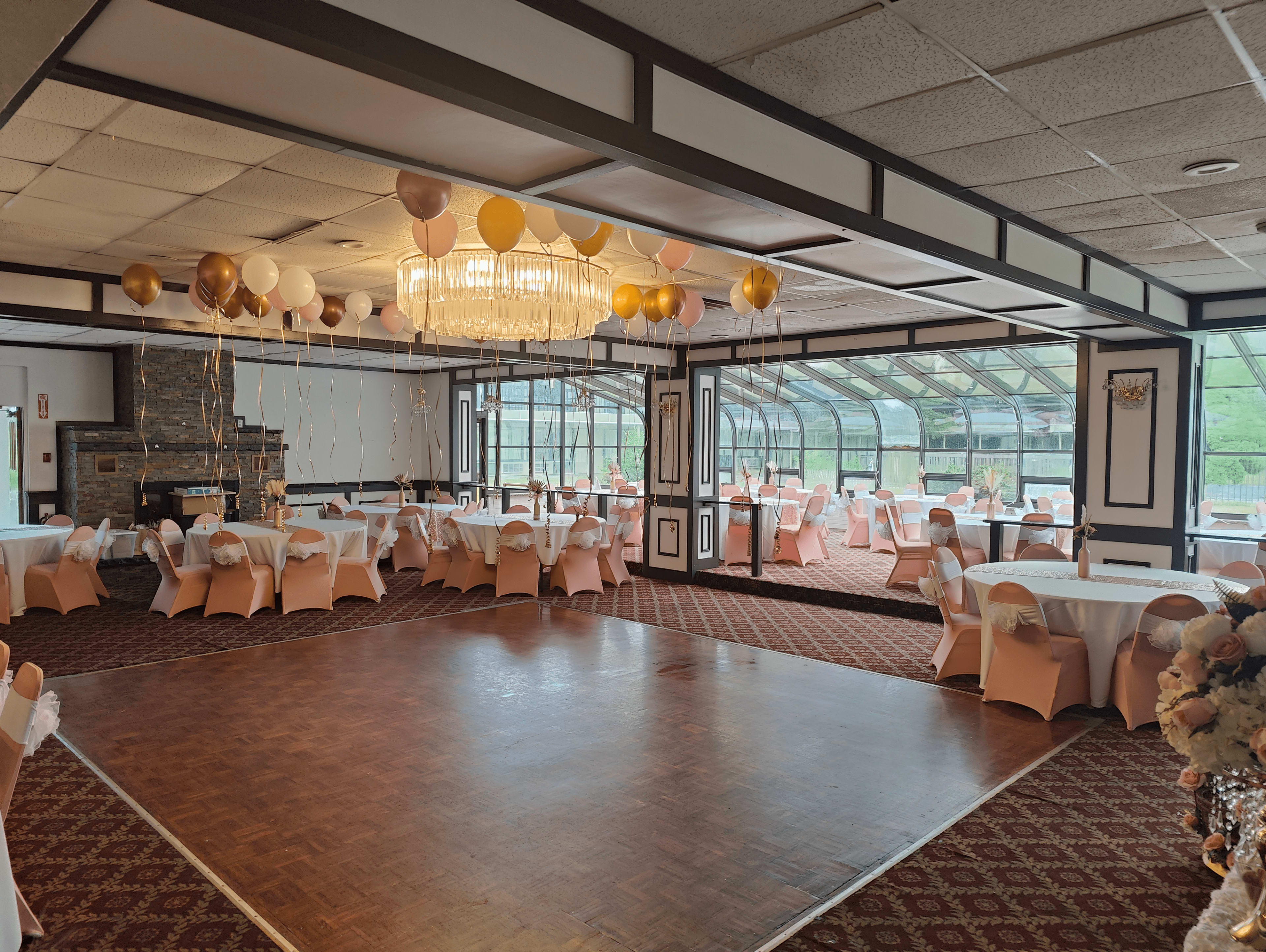A banquet hall with tables dressed in white and light pink linens, decorated with balloons and a chandelier, overlooking a glassed-in area.