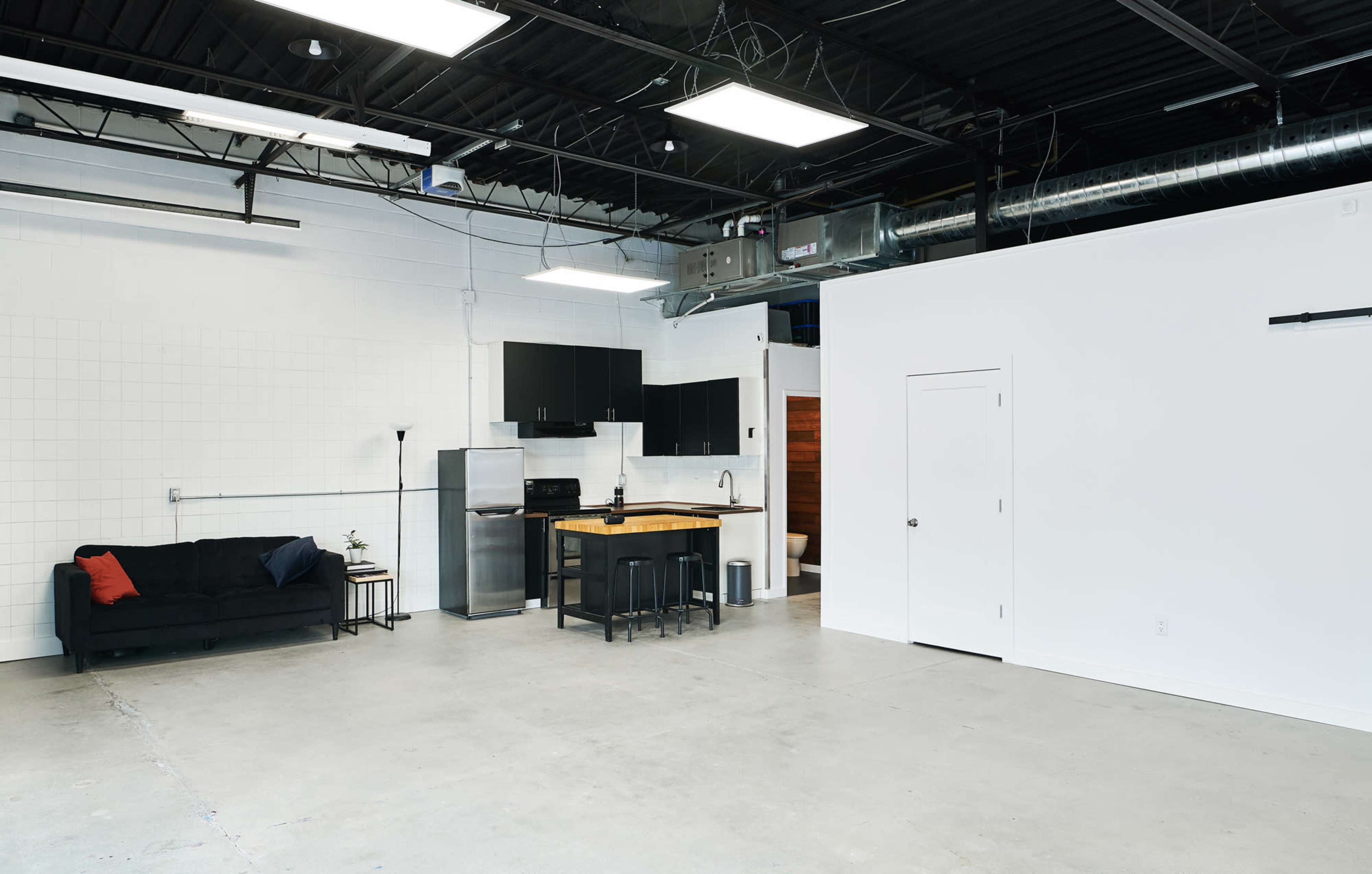 A minimalistic kitchen and living area with a black couch, a small dining table, and stainless steel appliances in an industrial-style space.