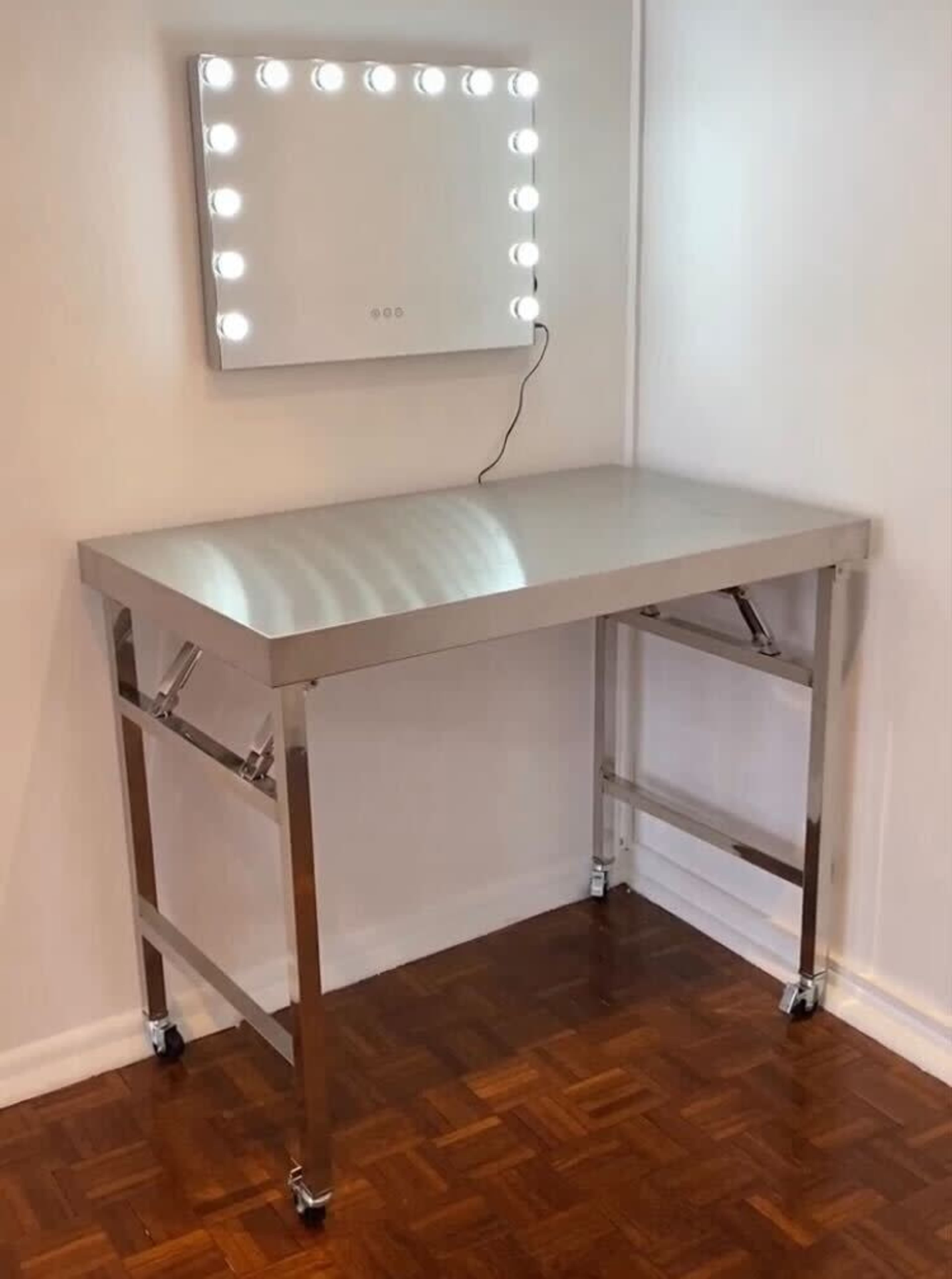 A modern, stainless steel table with a mirror illuminated by light bulbs, positioned against a wall in a room with wooden flooring.