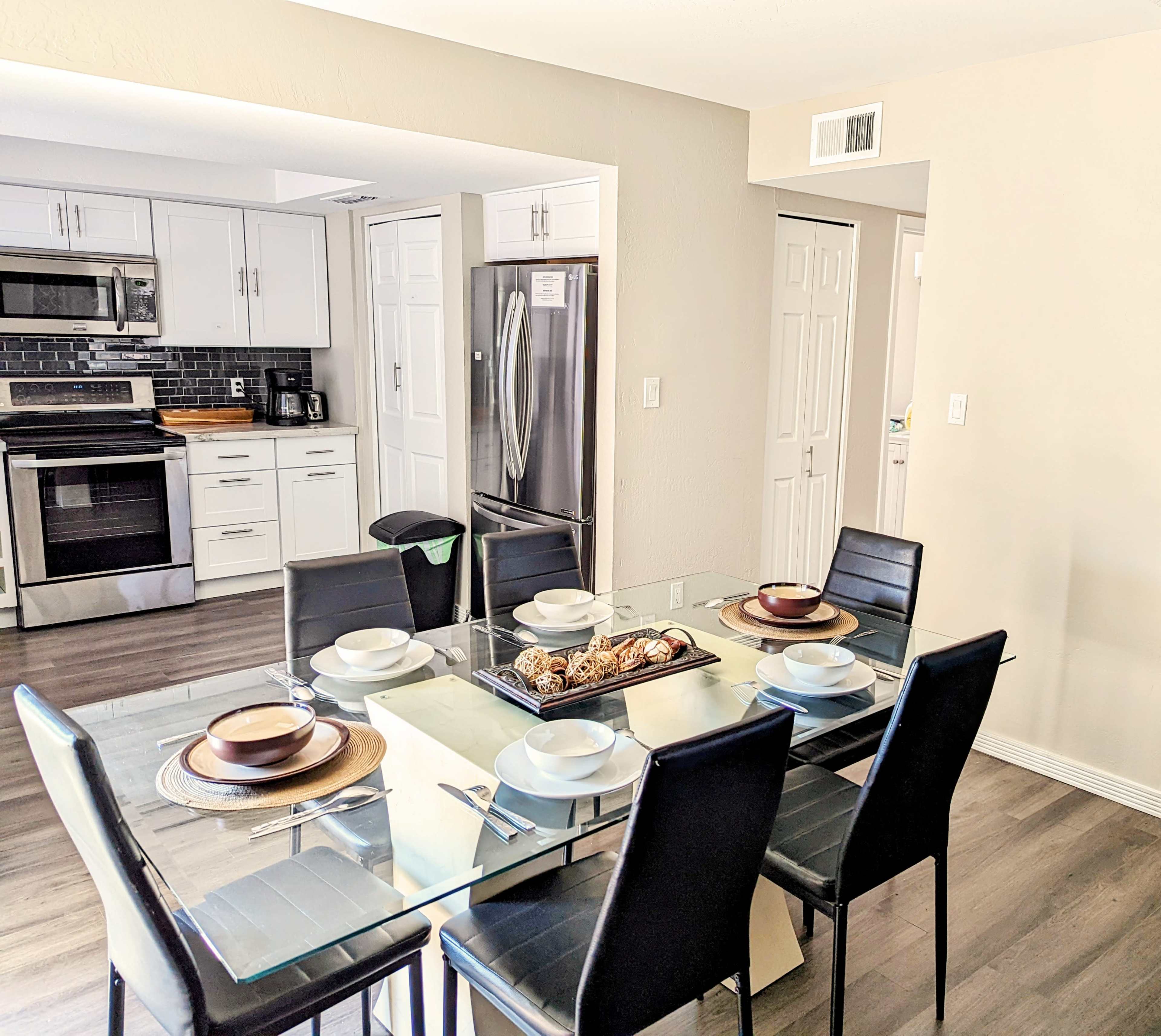 A modern dining room with a glass table set for six and a kitchen visible in the background.
