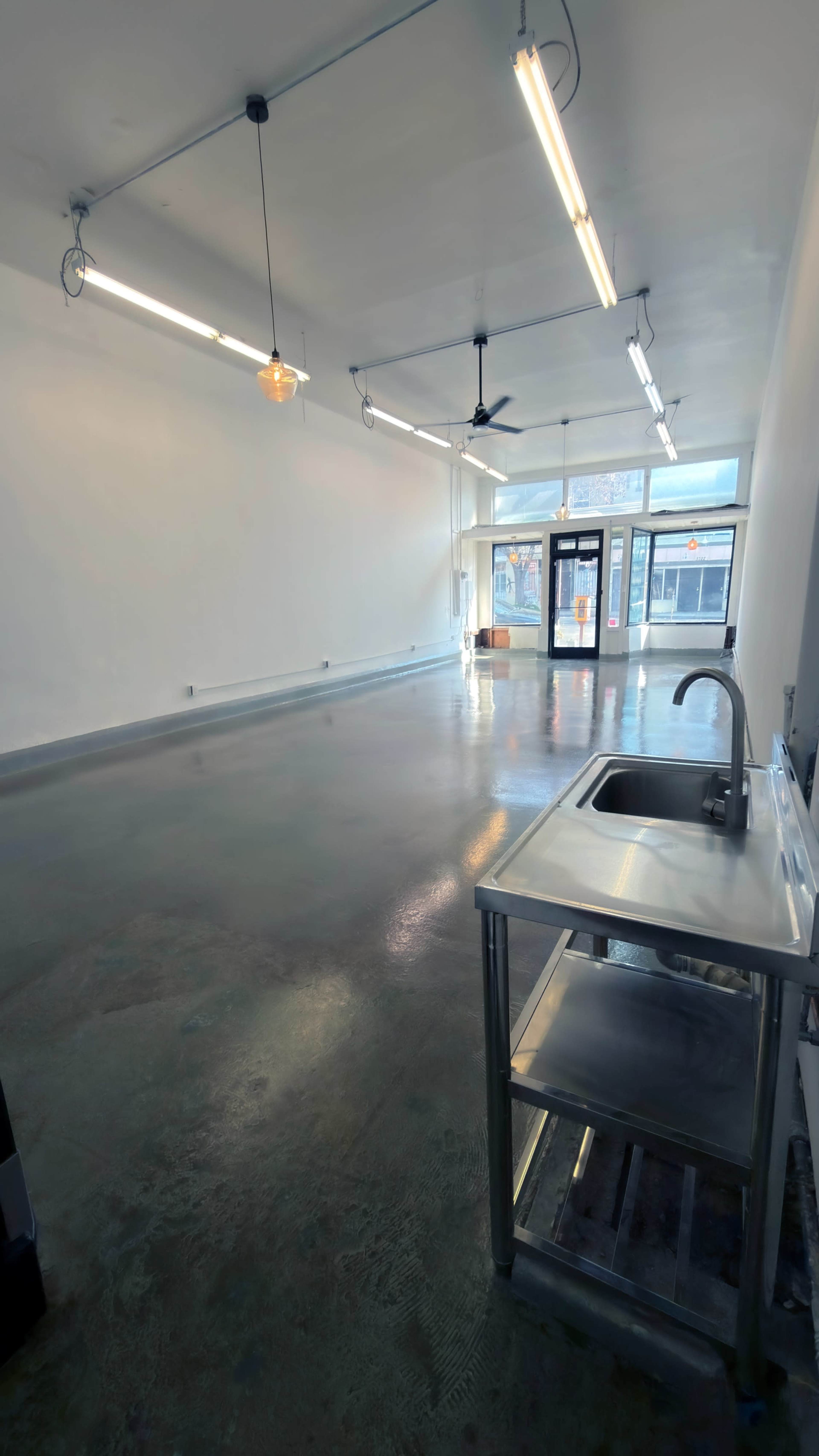 The image shows a spacious, empty commercial space with a sink on a stainless steel cart, large windows at the entrance, and fluorescent lighting overhead.