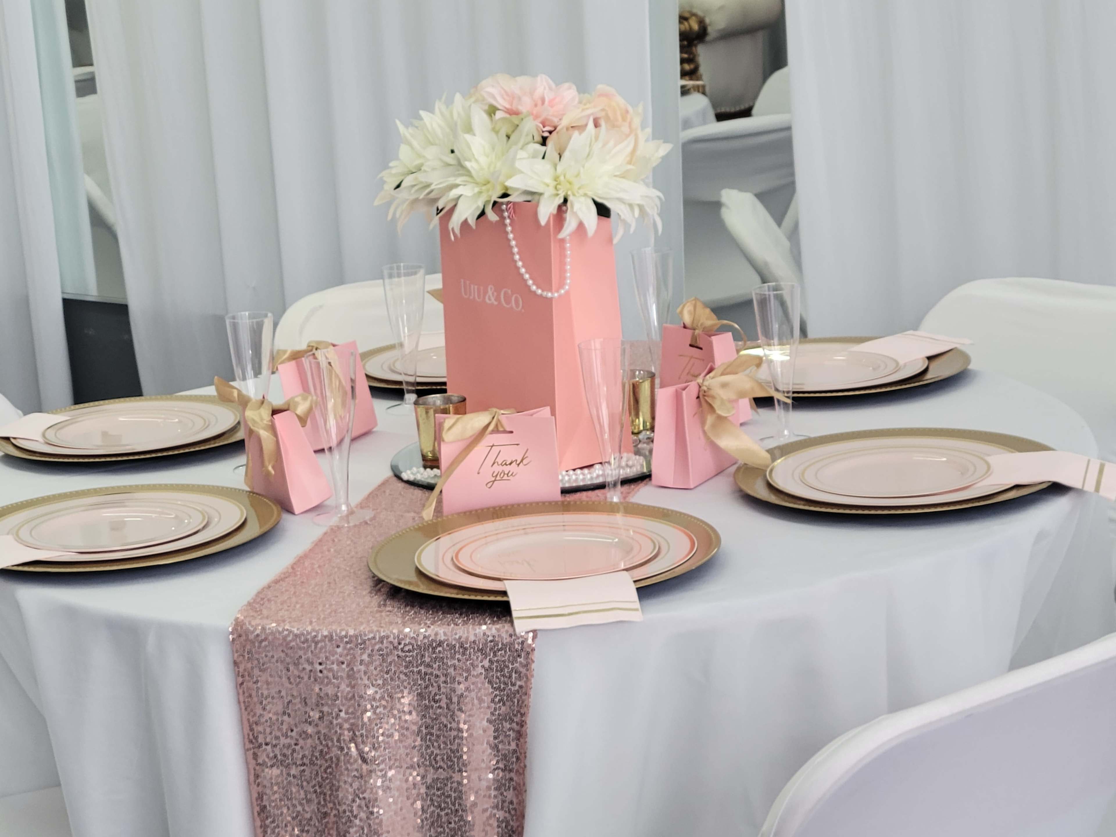 A dining table set with white and gold plates, pink favor bags, and a floral centerpiece arranged in a pink box.