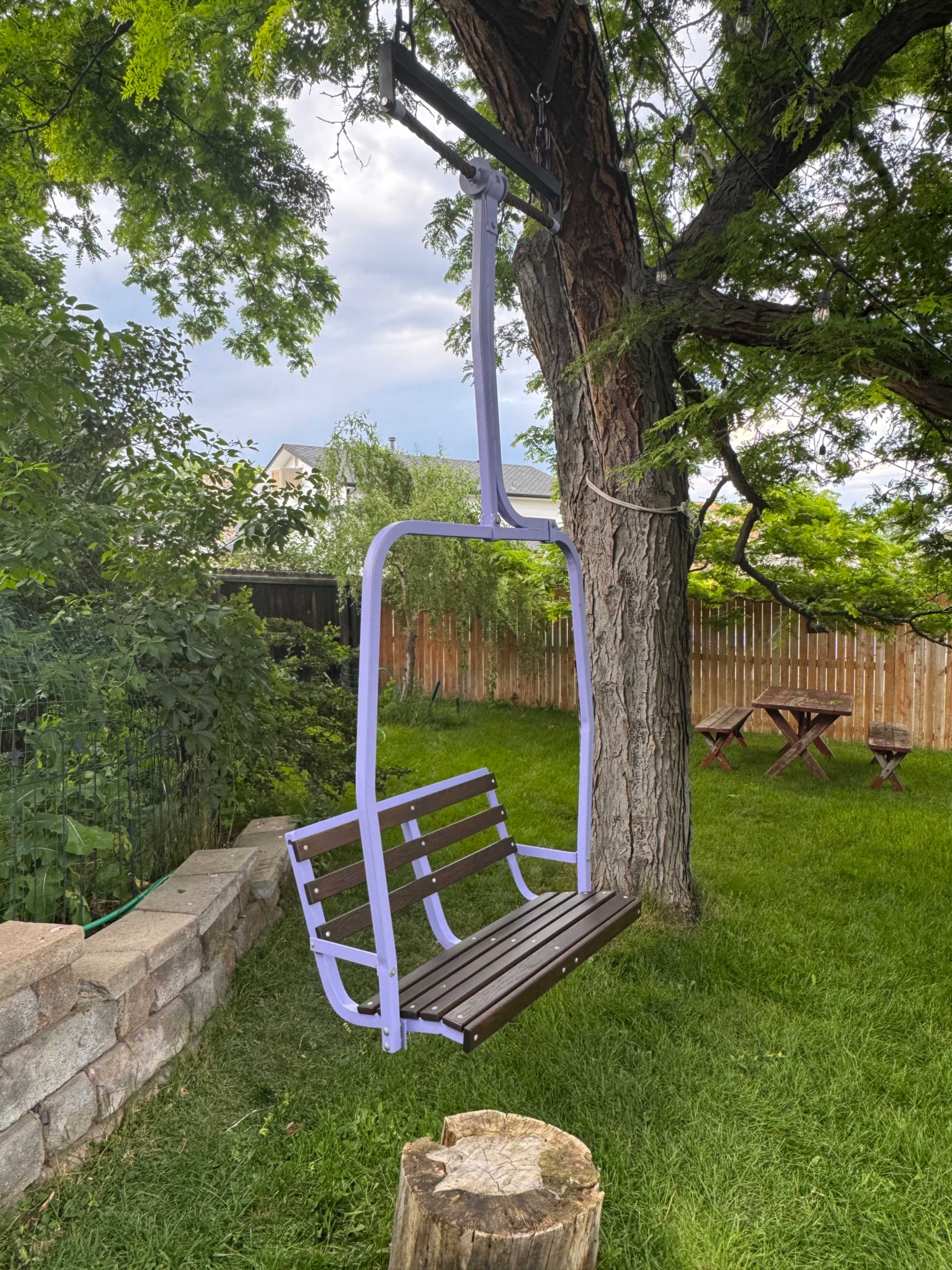 A chairlift-style swing is mounted on a tree in a backyard surrounded by greenery and wooden fencing.
