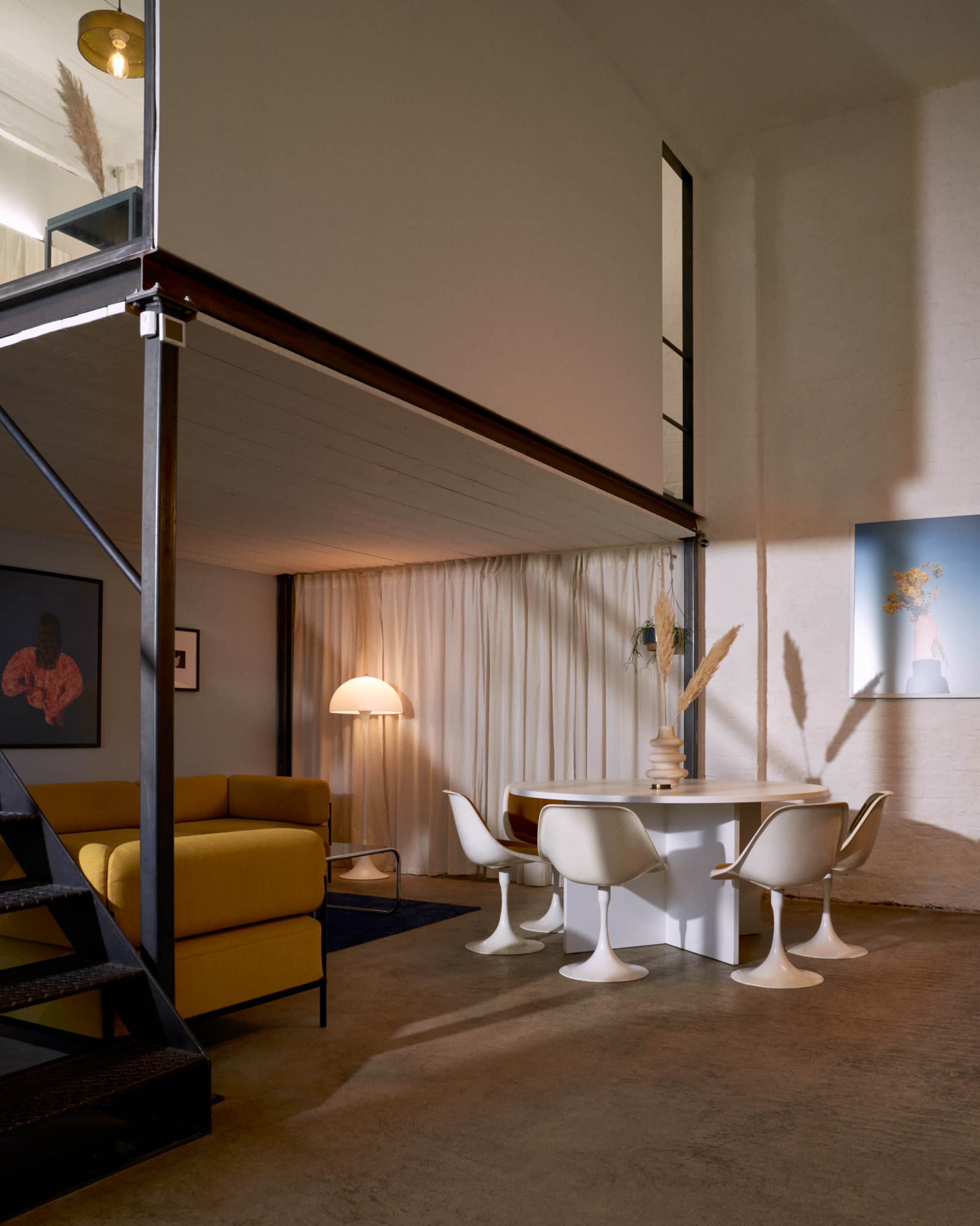 The image shows a modern interior featuring a yellow sofa, a circular white dining table with four white chairs, and a staircase leading to an upper level with large windows.