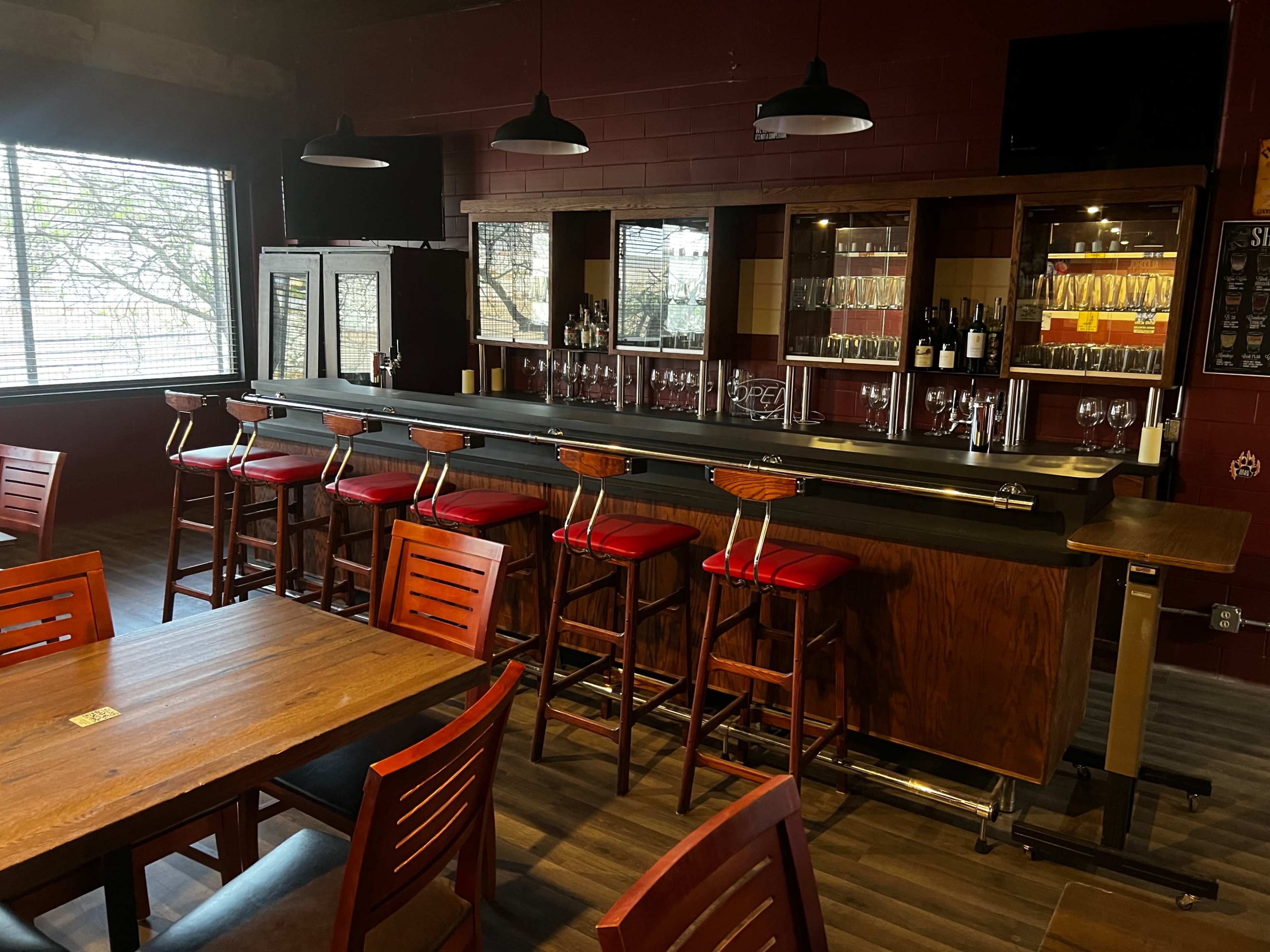 A bar area with a wooden counter, several high stools with red cushions, and shelves displaying bottles and glasses in a dimly lit establishment.