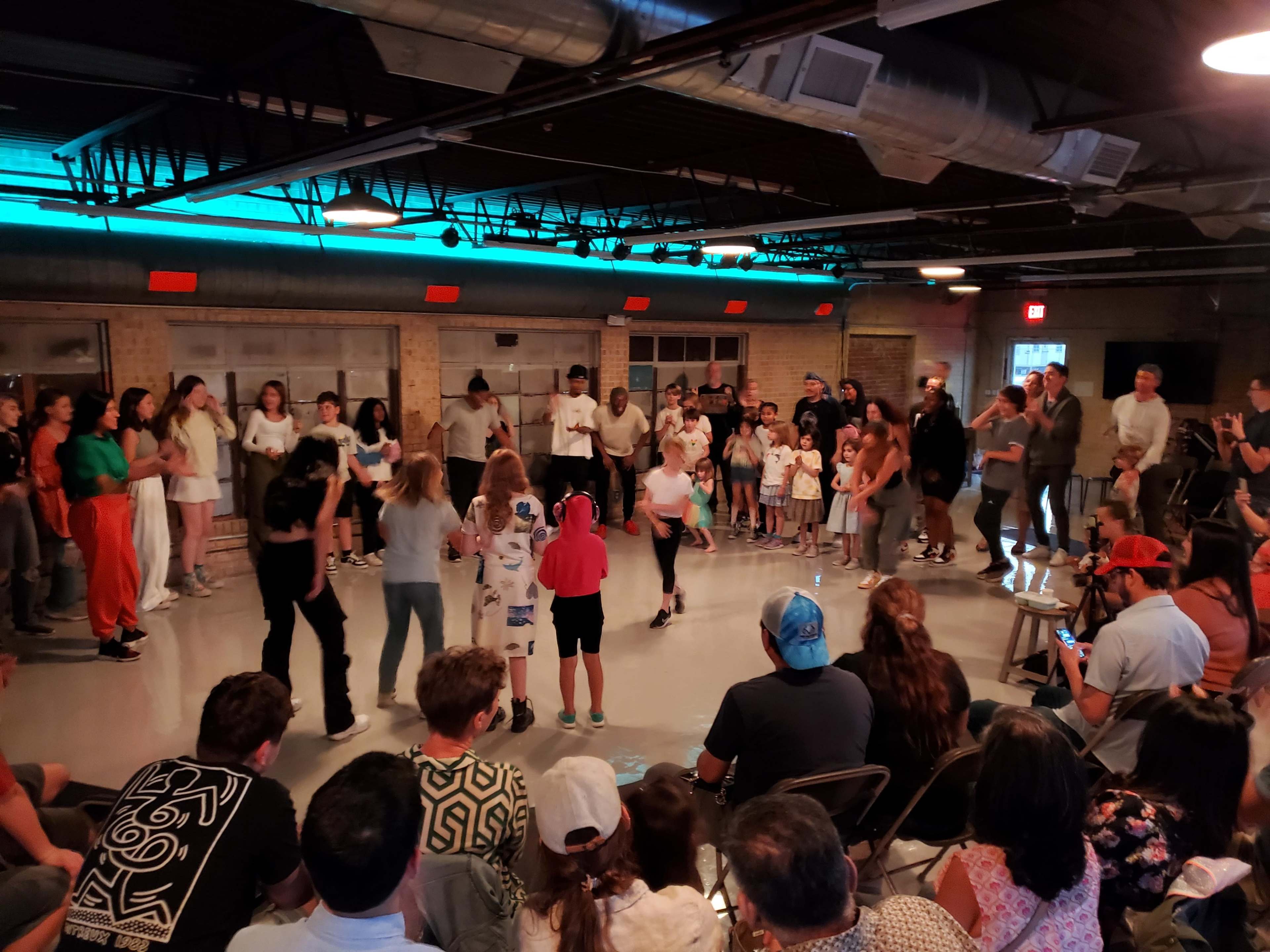A diverse group of people gathers in a spacious indoor venue, watching and participating in a lively dance performance.