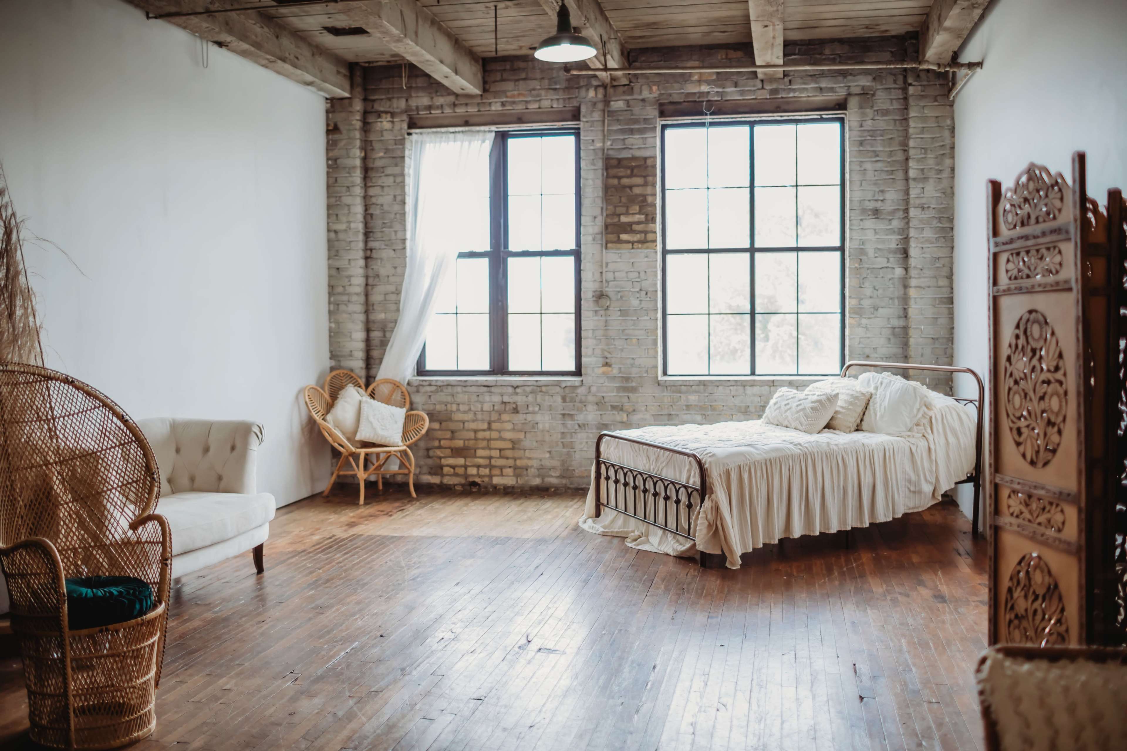 A spacious bedroom with large windows, a metal bed with a white bedspread, and wicker furniture arranged around a wooden floor.