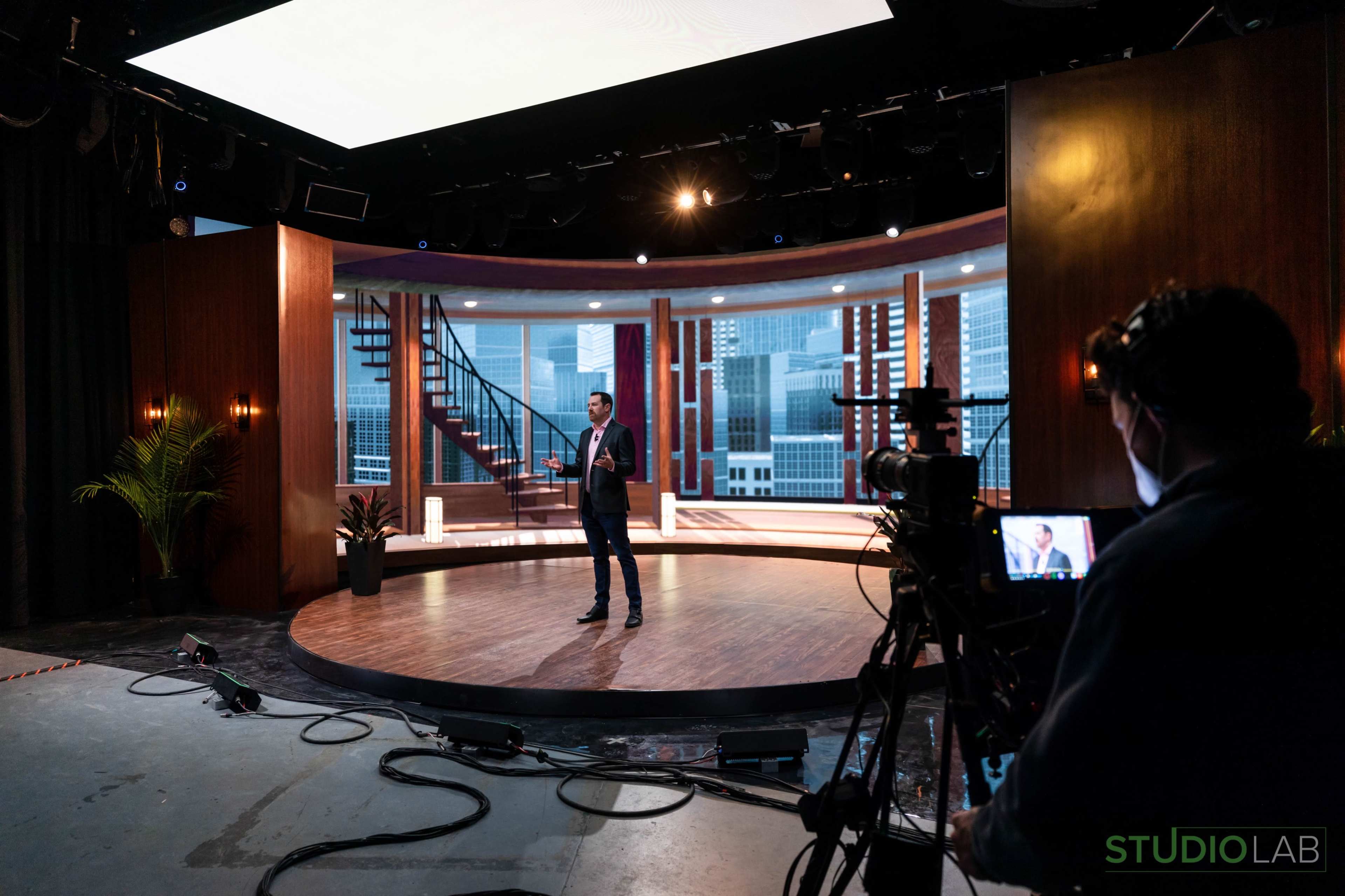 A man stands on a stage with a modern backdrop featuring large windows and a staircase, while a camera operator captures the scene.