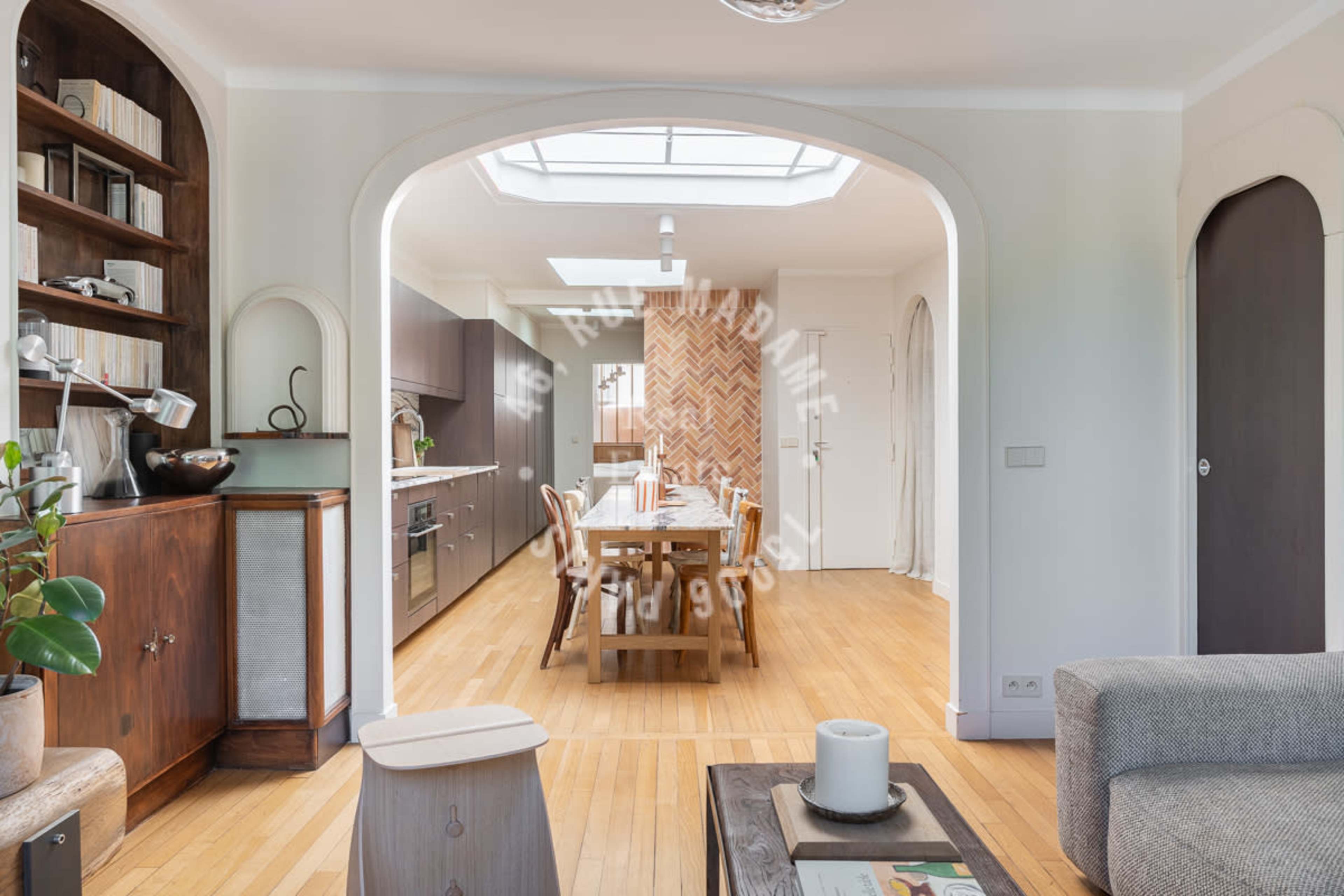 The image shows a modern kitchen and dining area with wooden flooring, an island counter, and a long dining table under a skylight.