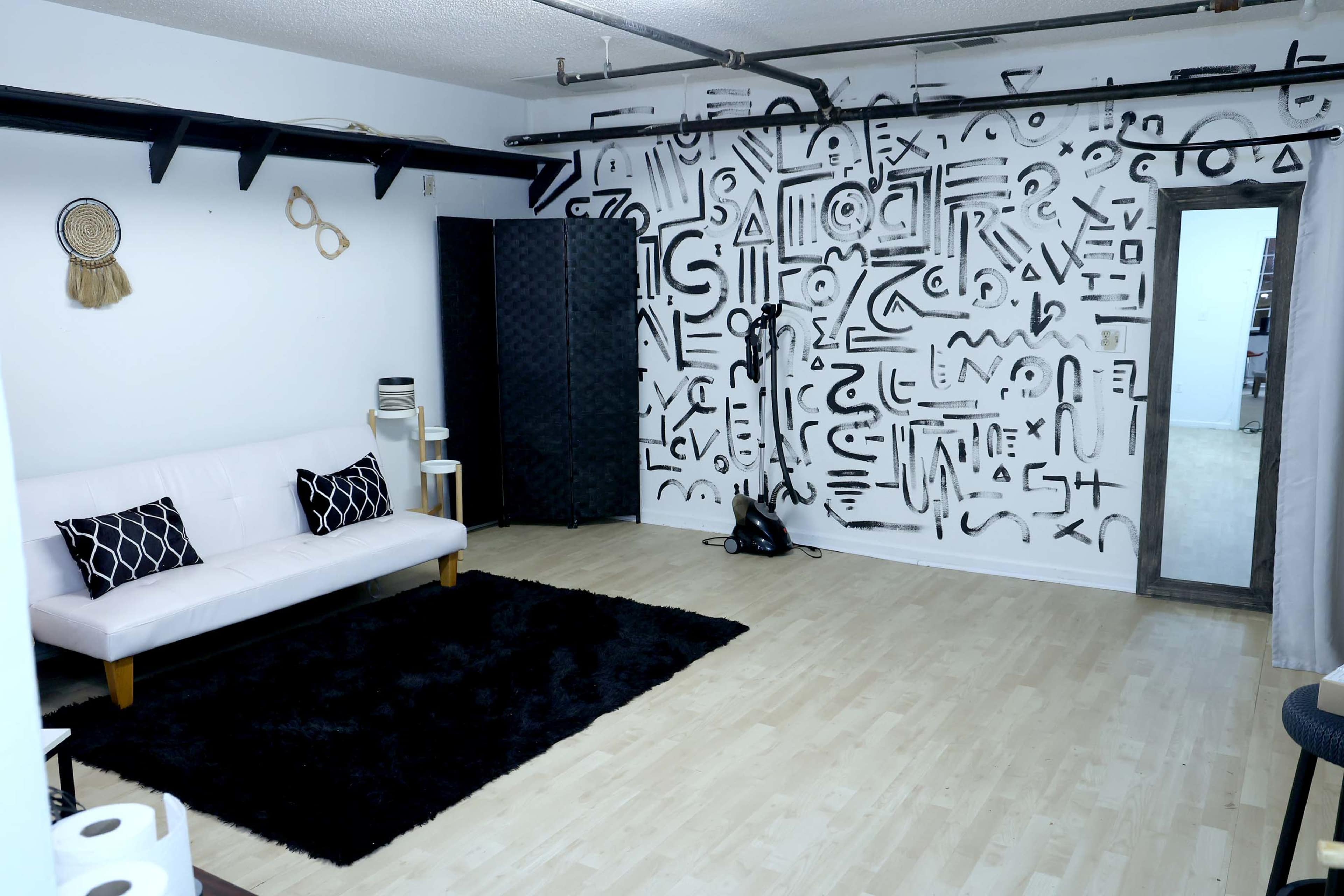 The image shows a minimalist indoor space featuring a white sofa, a black rug, decorative wall art, and a partially visible door leading to another room.