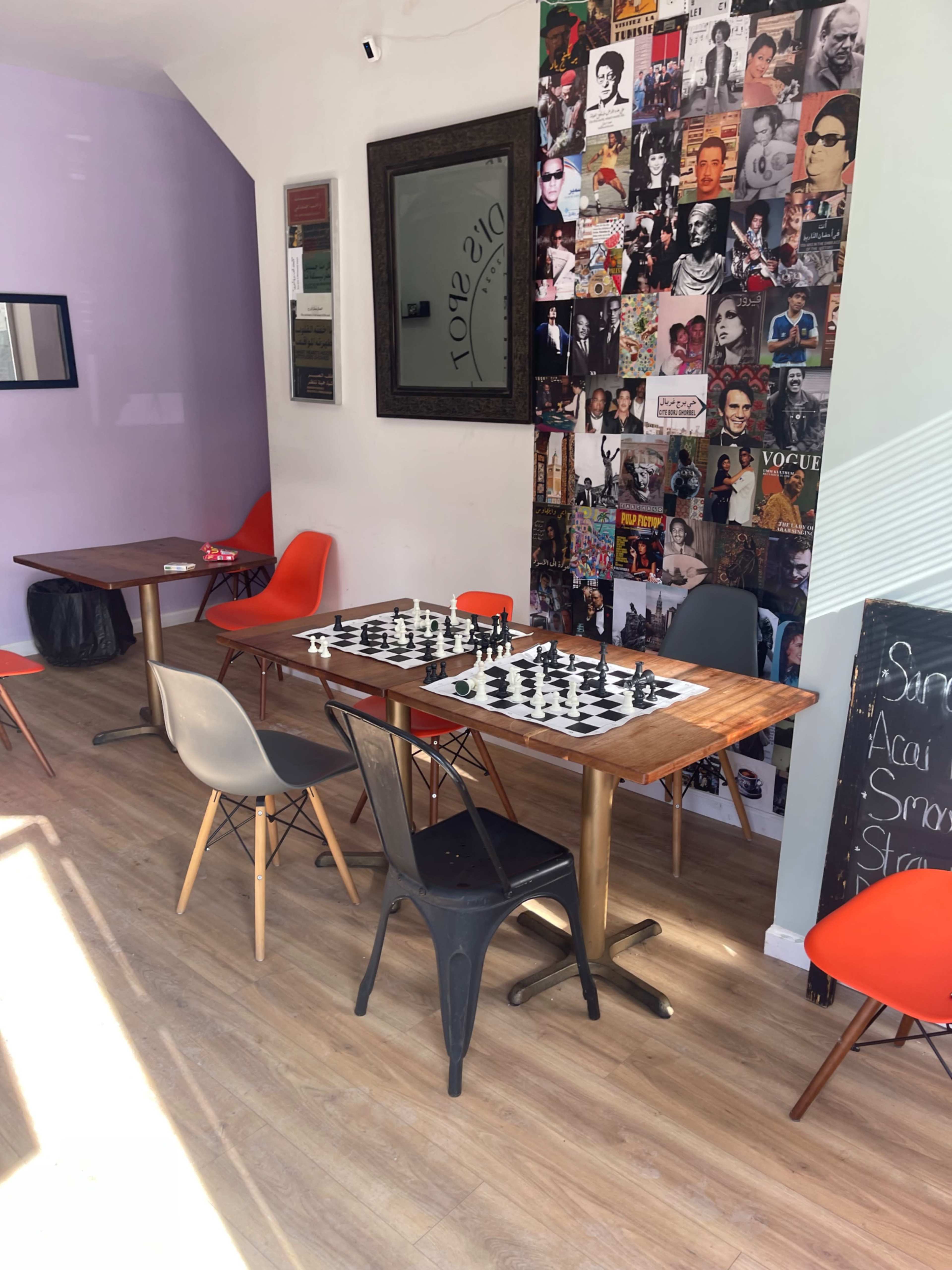 The image shows a cozy cafe with two wooden tables surrounded by colorful chairs, featuring a large wall adorned with black-and-white photographs and a chessboard set up on one of the tables.