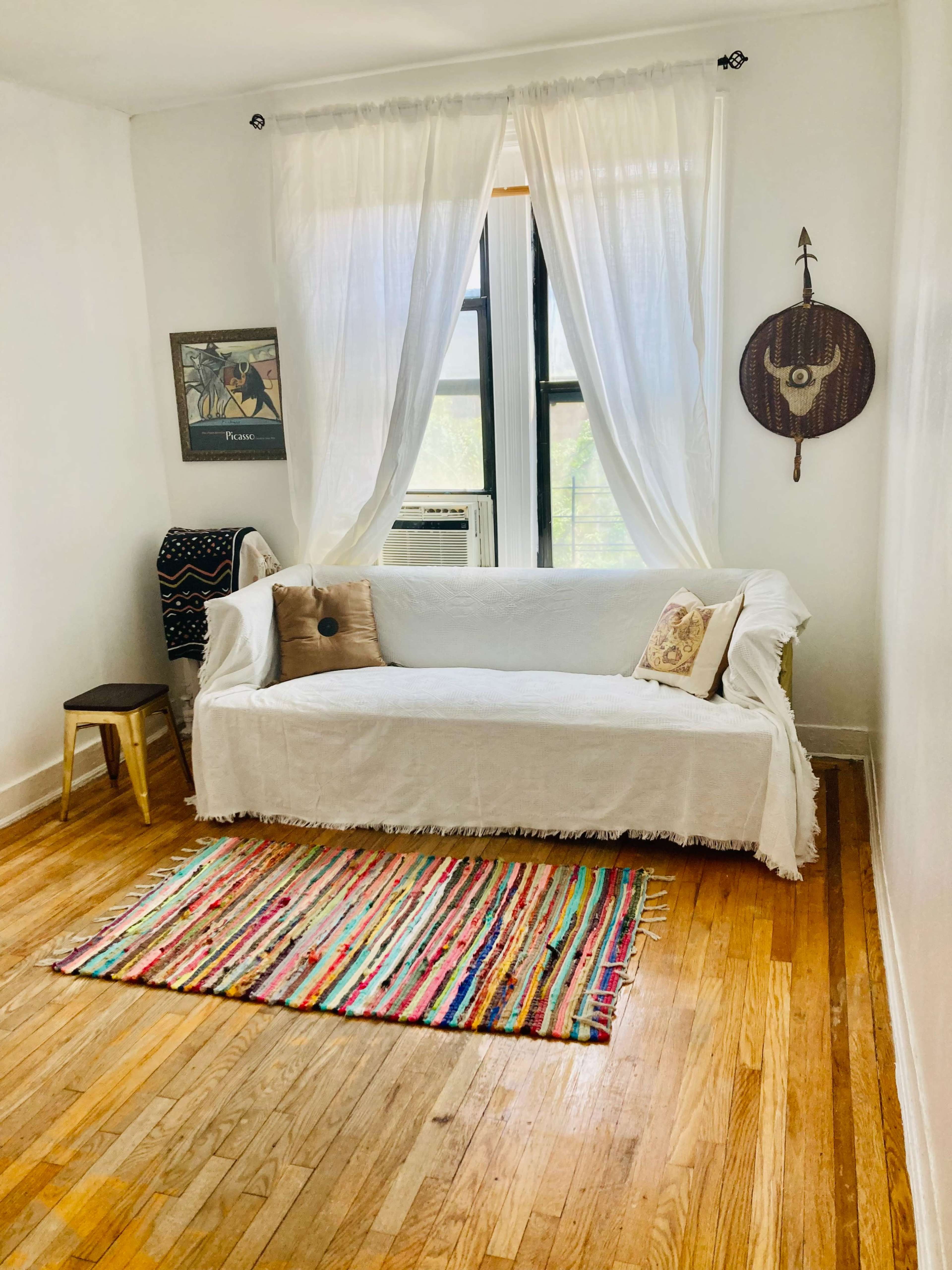 A bright, minimalistic living room featuring a white sofa covered with a throw, a woven rug, wooden flooring, and large windows adorned with sheer curtains.