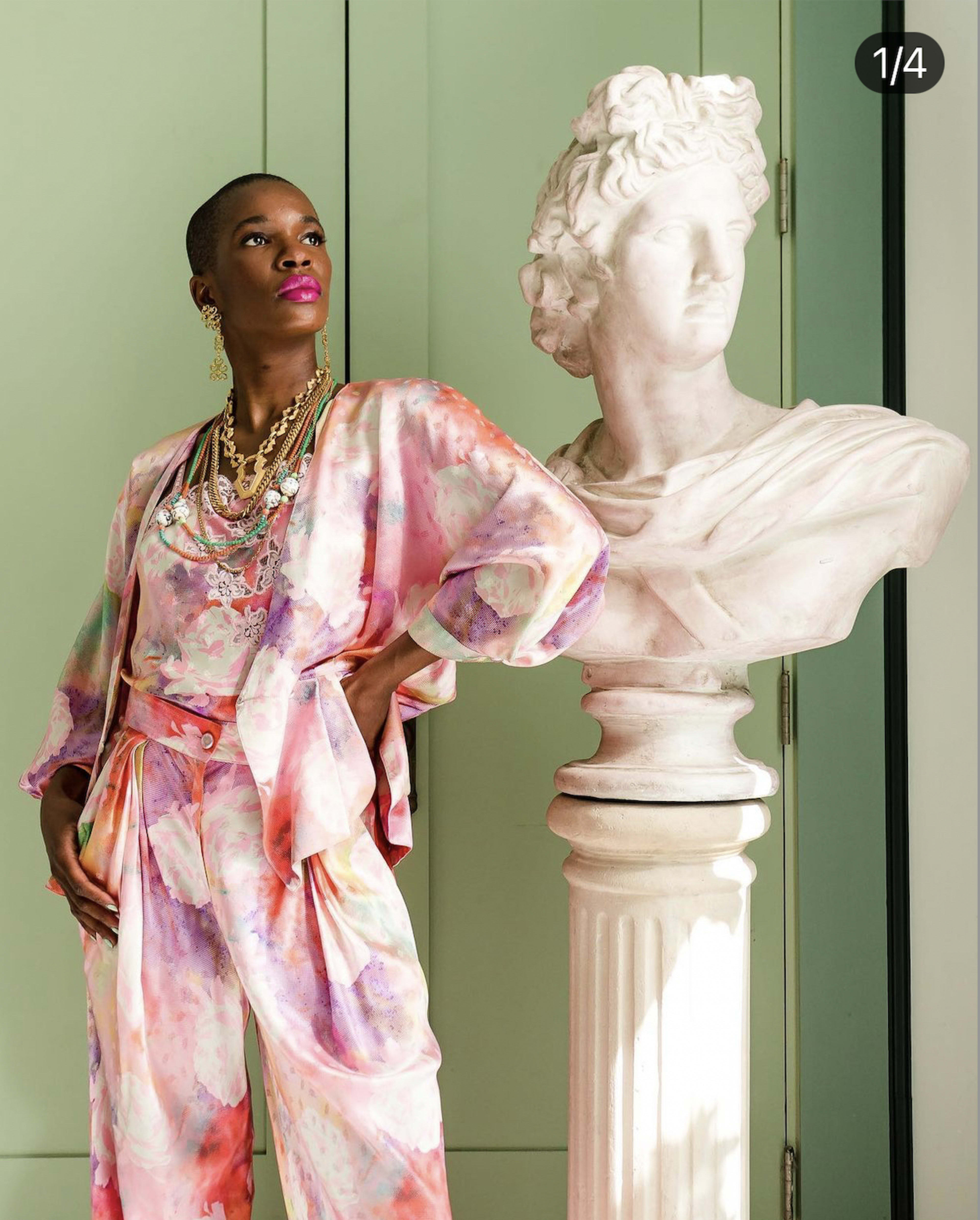 A person in a colorful outfit poses next to a marble bust in a well-lit room with mint green walls.