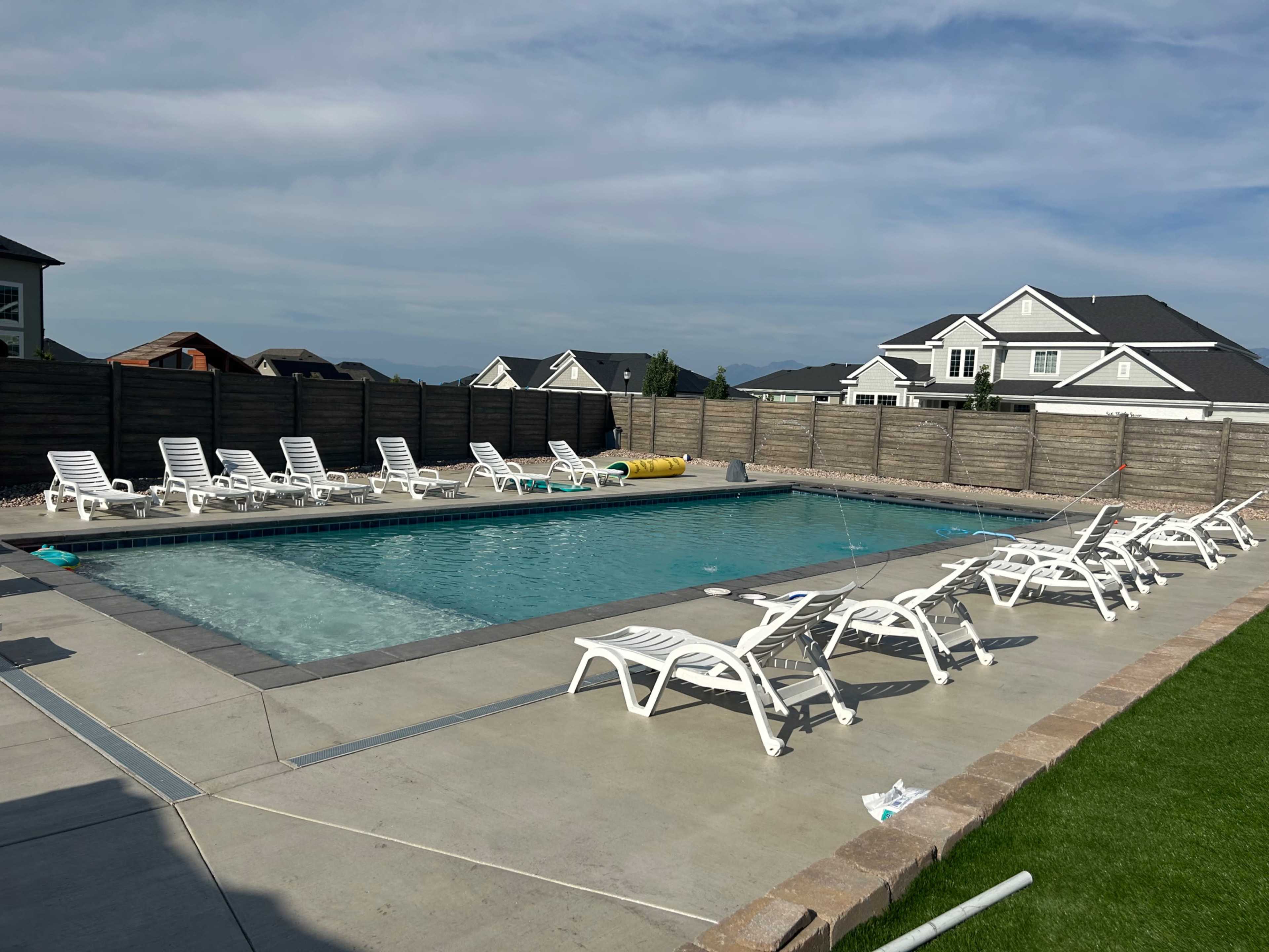 A rectangular swimming pool is surrounded by lounge chairs on a concrete deck, with a wooden fence and houses visible in the background.