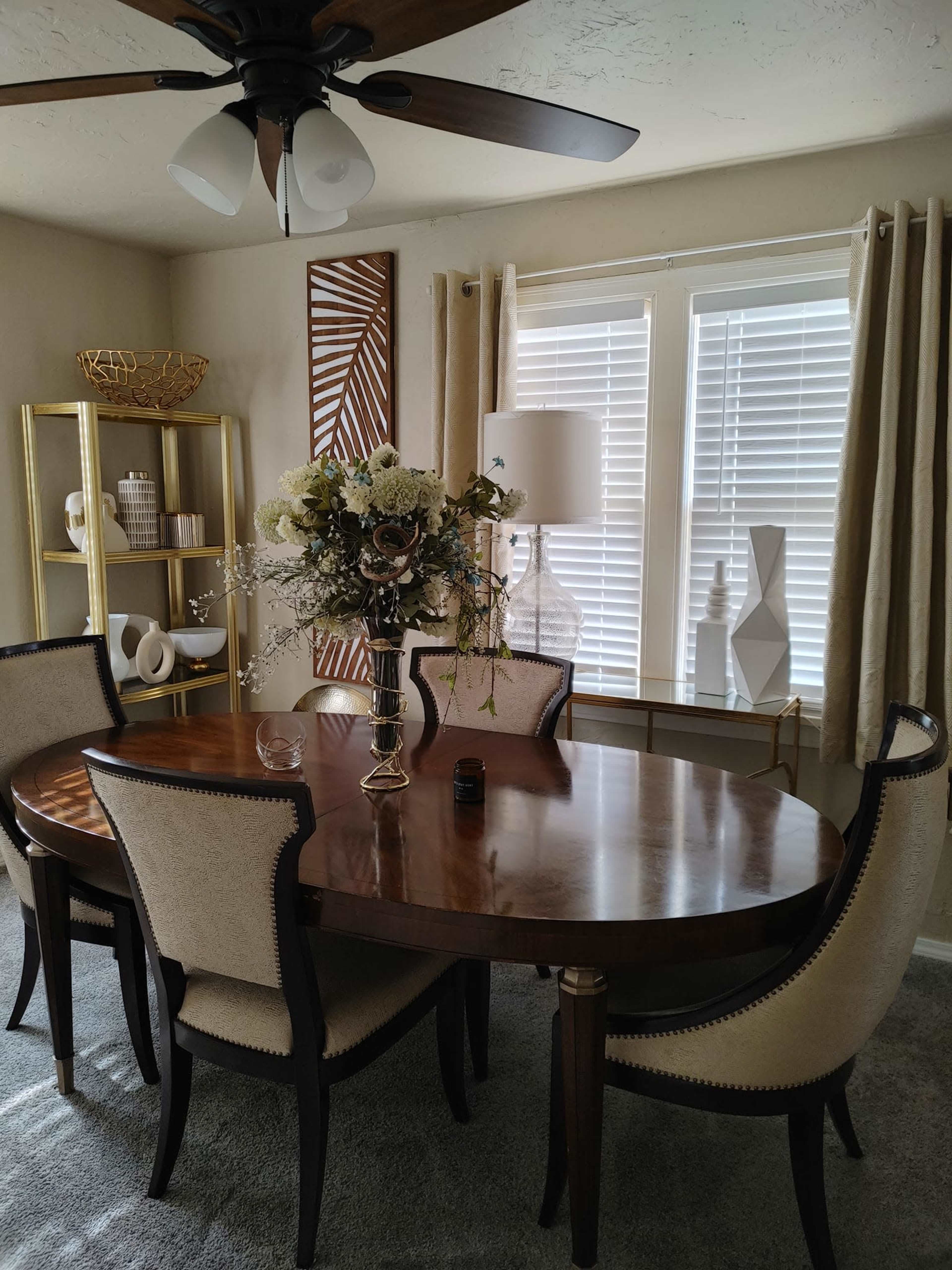 A wooden dining table surrounded by upholstered chairs is set near large windows adorned with closed blinds and decorative furnishings.