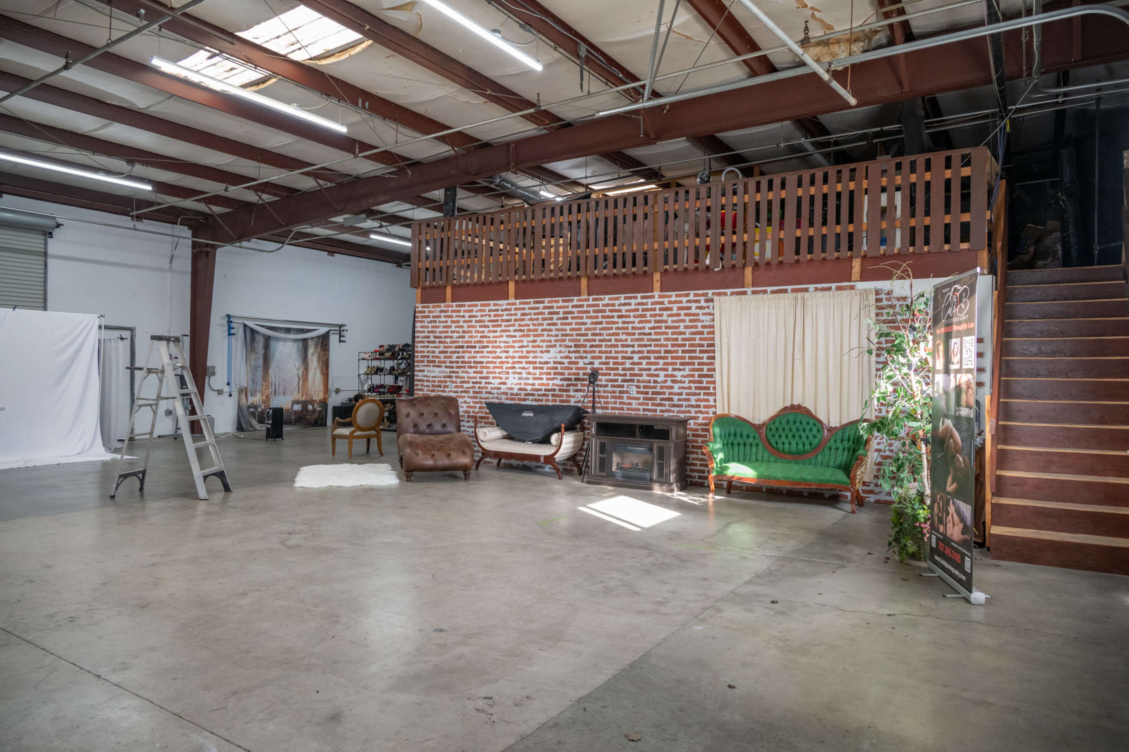 The image shows a spacious warehouse interior with exposed steel beams, a lofted area, and a mix of seating options, including vintage couches, a ladder, and a backdrop for photography.