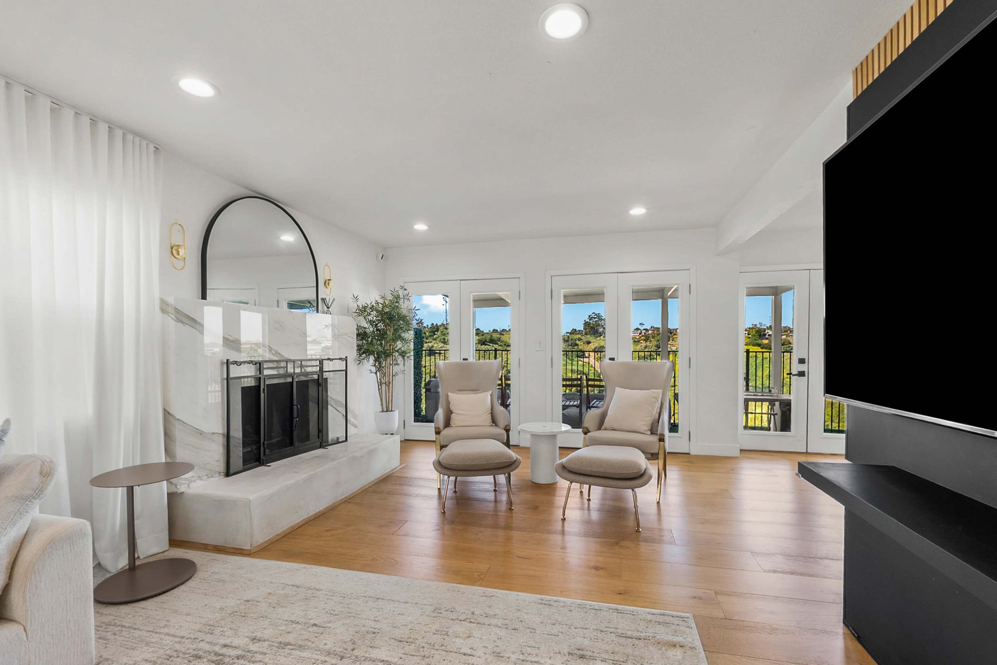 The image shows a modern living room with two armchairs facing a marble fireplace, large windows opening to a view, and a mounted television on a black wall.