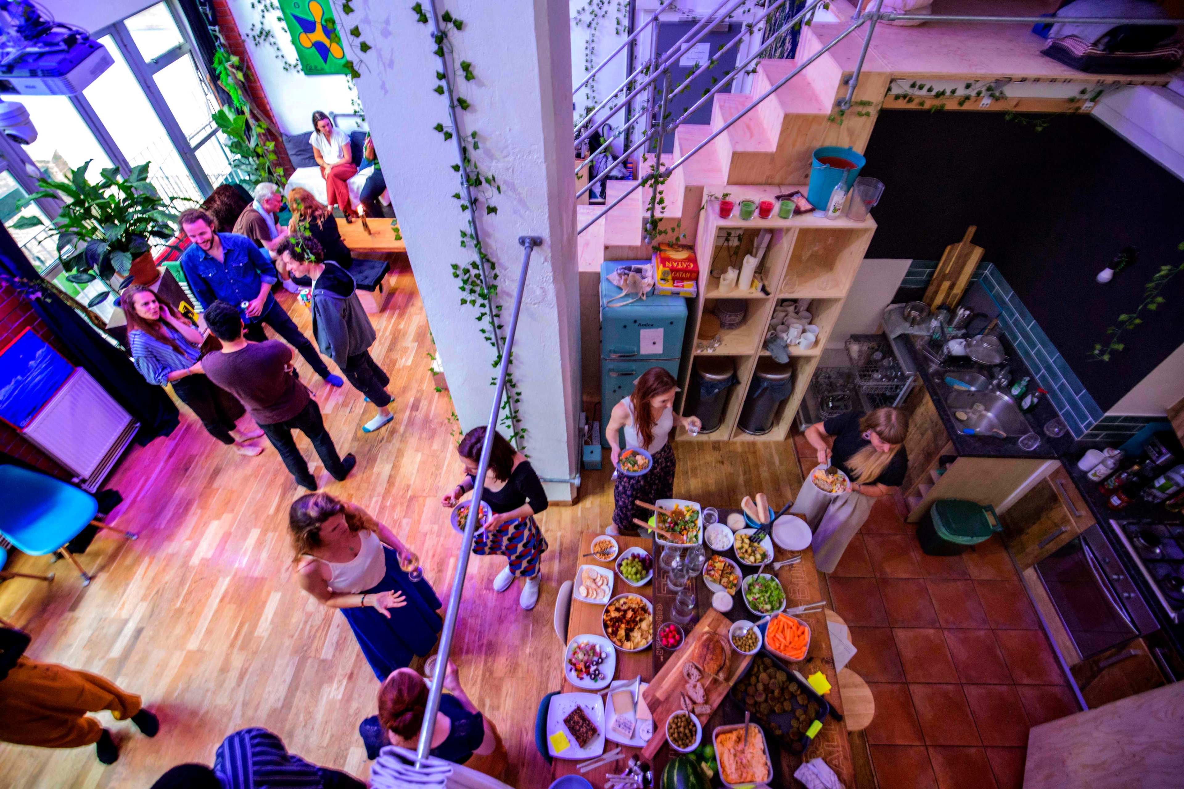 A lively gathering takes place in a spacious communal area with people socializing and a table full of various dishes.