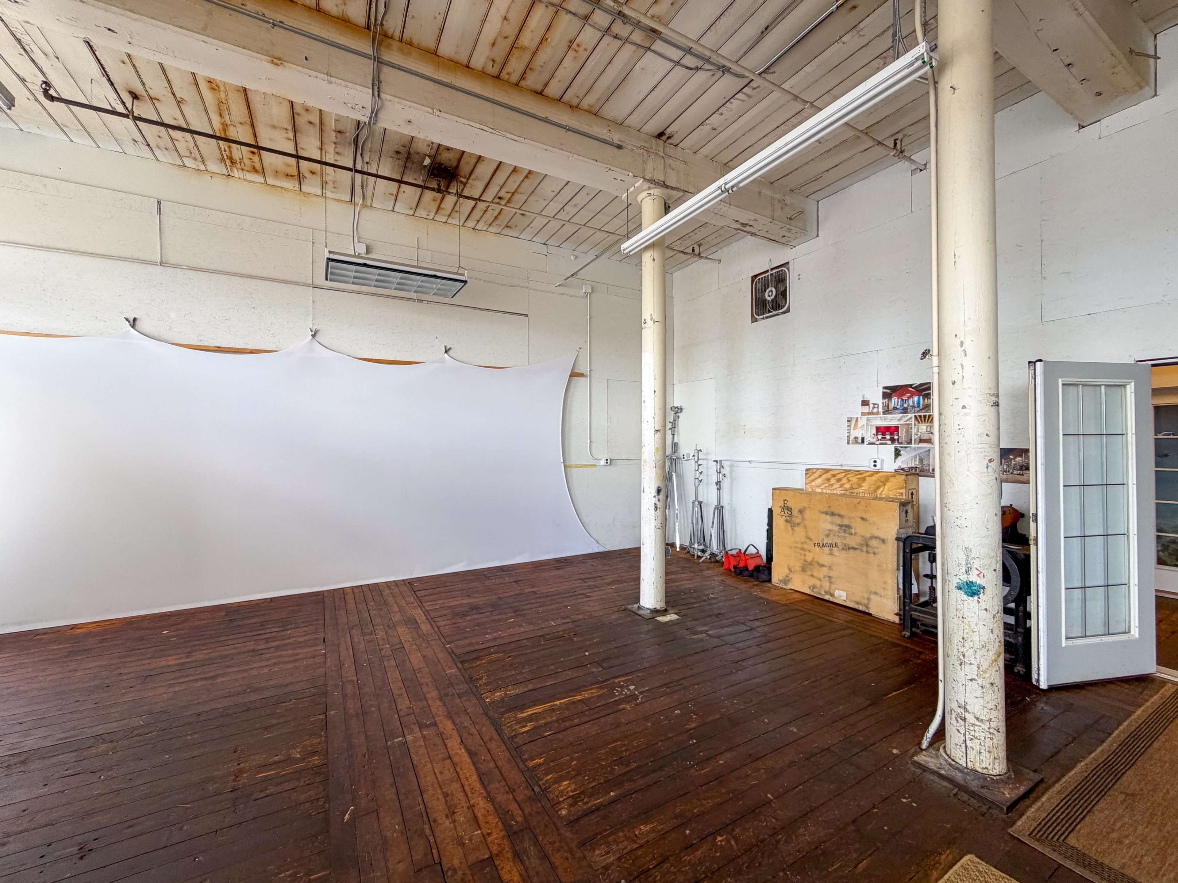 The image shows an empty studio space with a white backdrop, wooden flooring, and various equipment scattered along the walls.