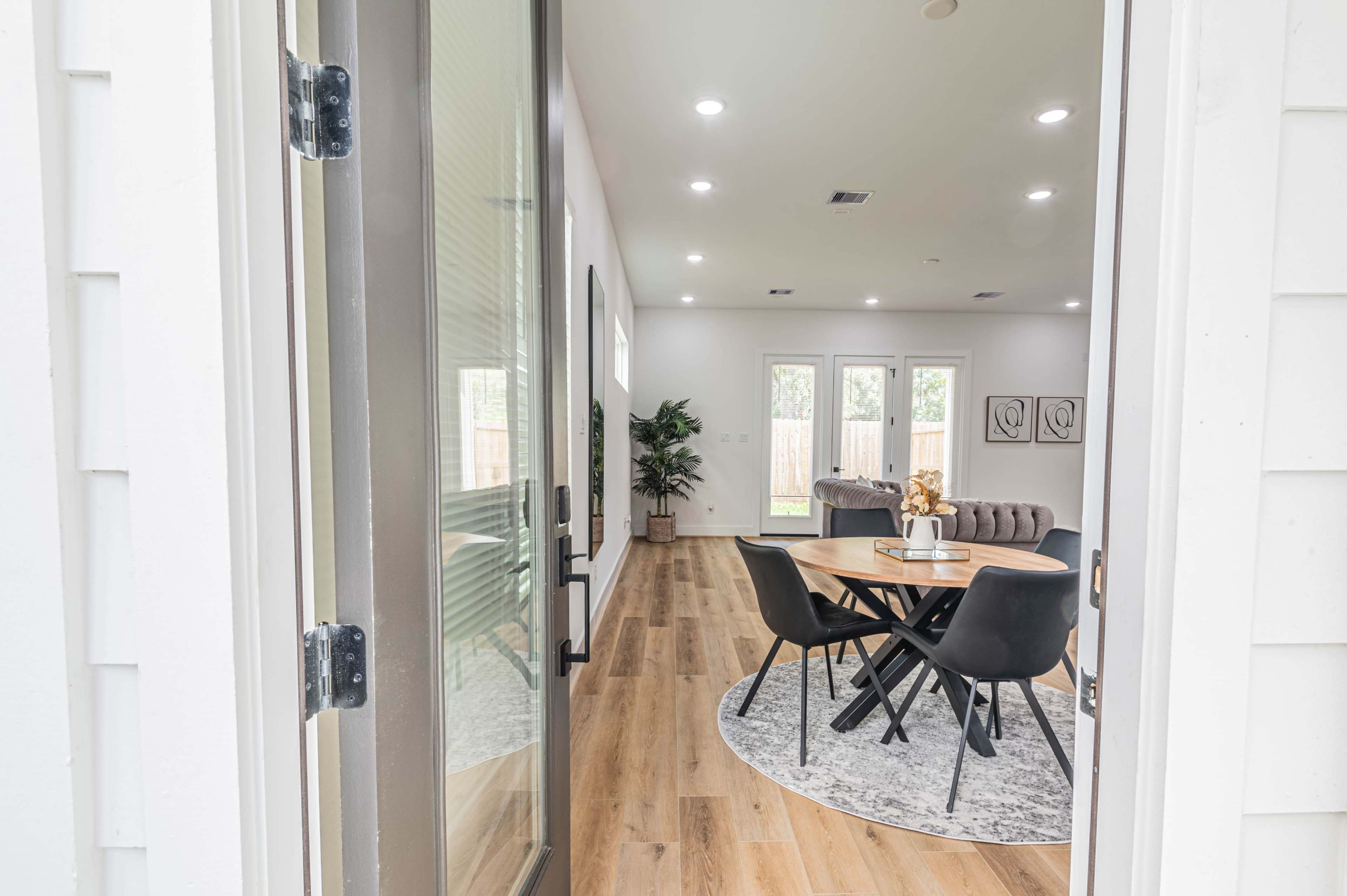 The interior of a modern home viewed from an open front door, featuring a circular dining table with black chairs and a living area in the background.
