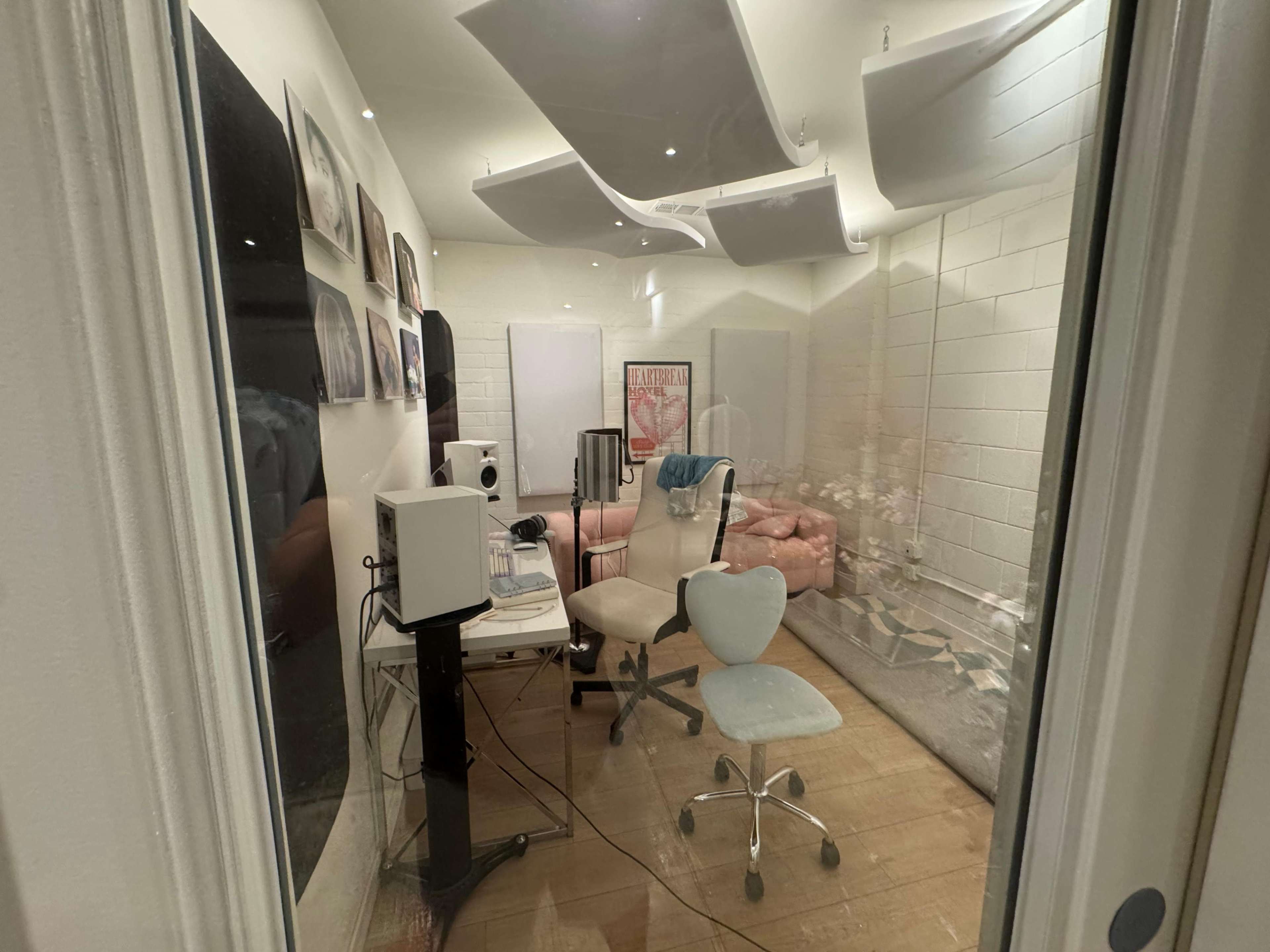 The image shows a small recording studio with a computer setup, two chairs, acoustic panels, and artwork on the walls.